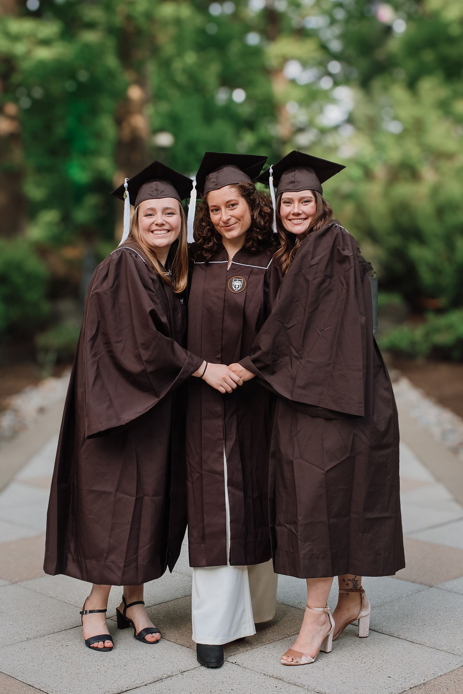 Three graduates in brown caps and gowns pose together on a stone pathway during commencement celebrations.