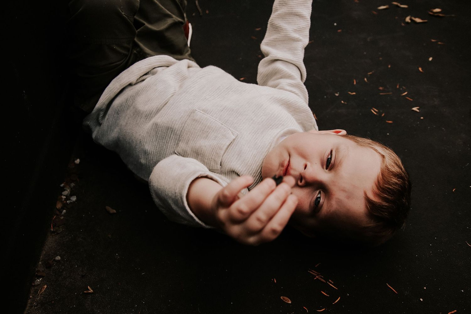 Person wearing white sweater lying on dark ground with scattered autumn leaves covering their face with hands.