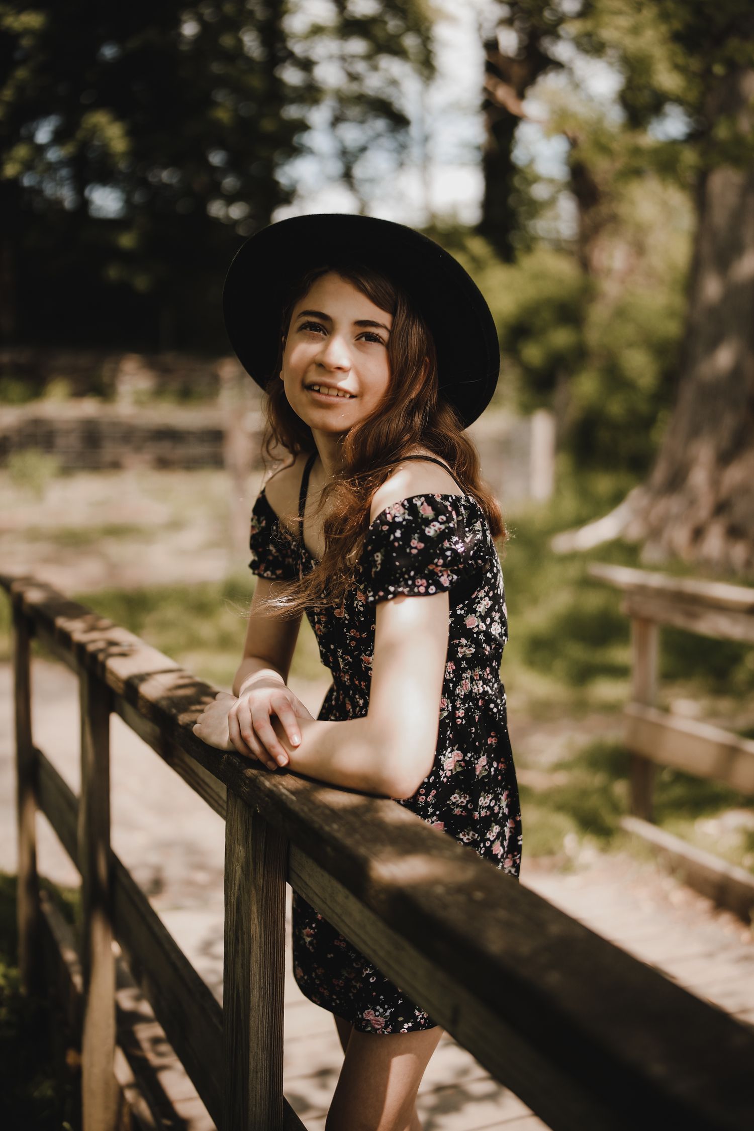 A person in a floral dress and black hat leans against a wooden railing in a sunlit forest setting.