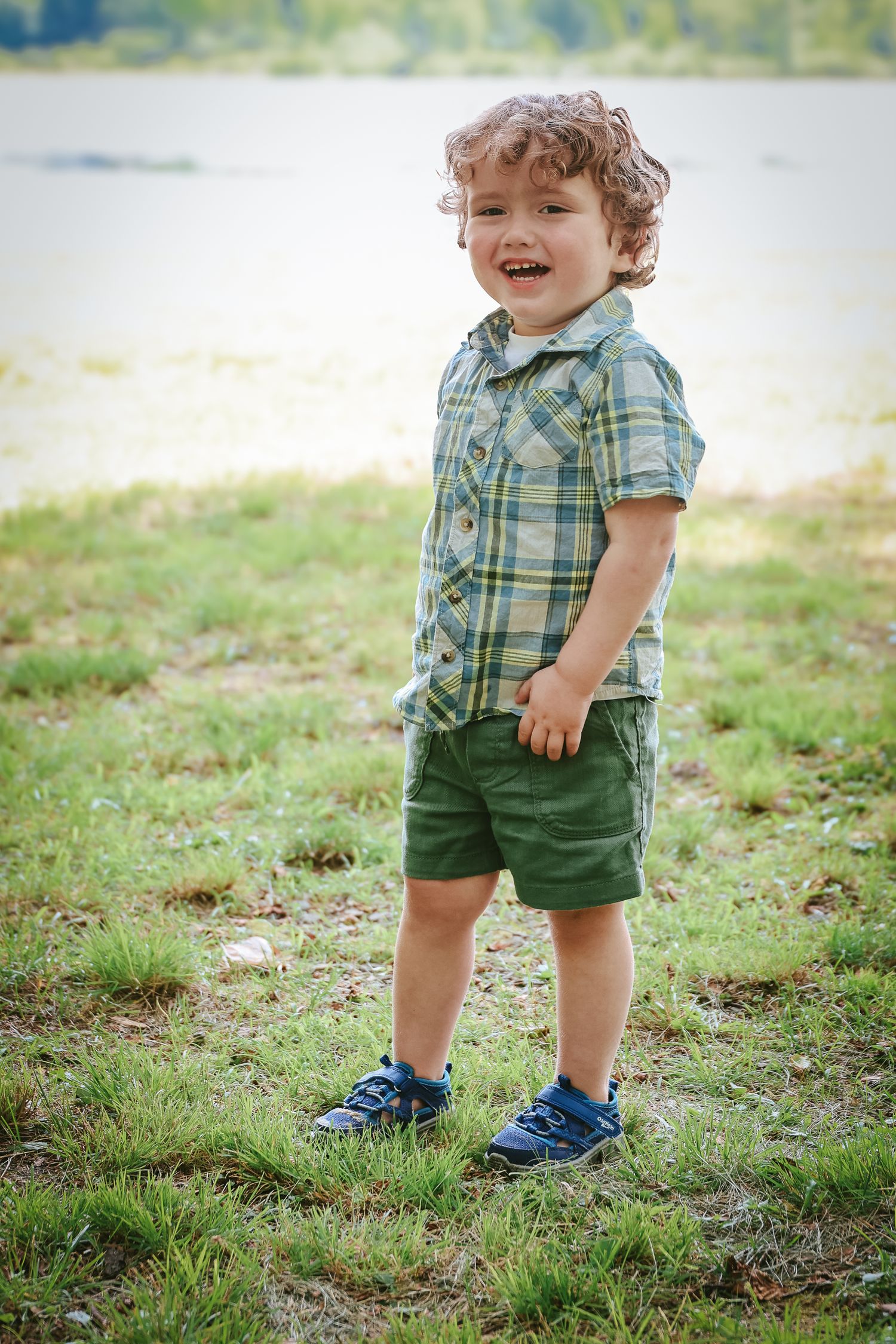 A child in plaid shirt and green shorts stands on grass wearing blue sneakers.