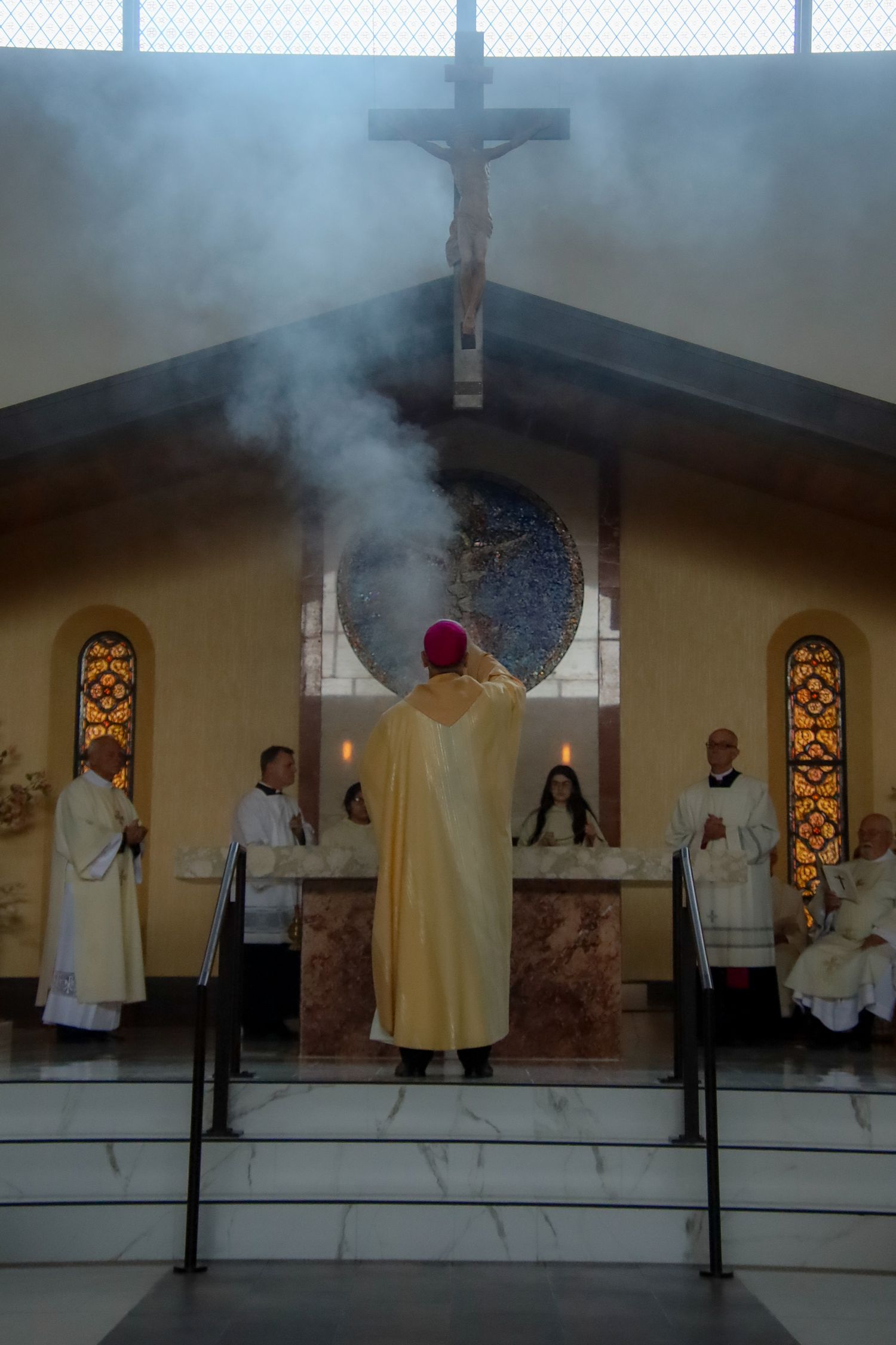 Religious ceremony with clergy in white and gold robes standing at altar amid rising incense smoke in church setting.