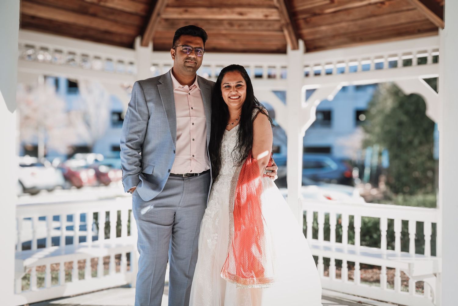 A couple in wedding attire stands under a white gazebo during a romantic outdoor ceremony.
