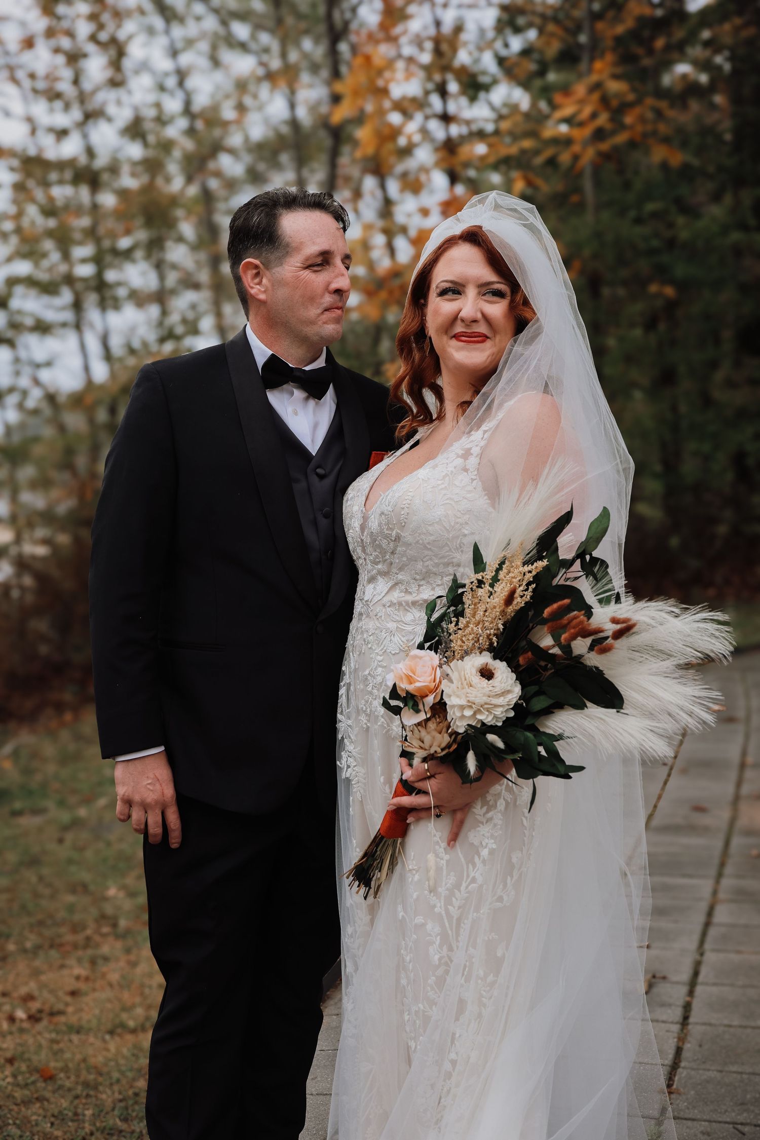 Newlywed couple shares romantic moments in autumn outdoor setting with fall foliage and white bridal bouquet.