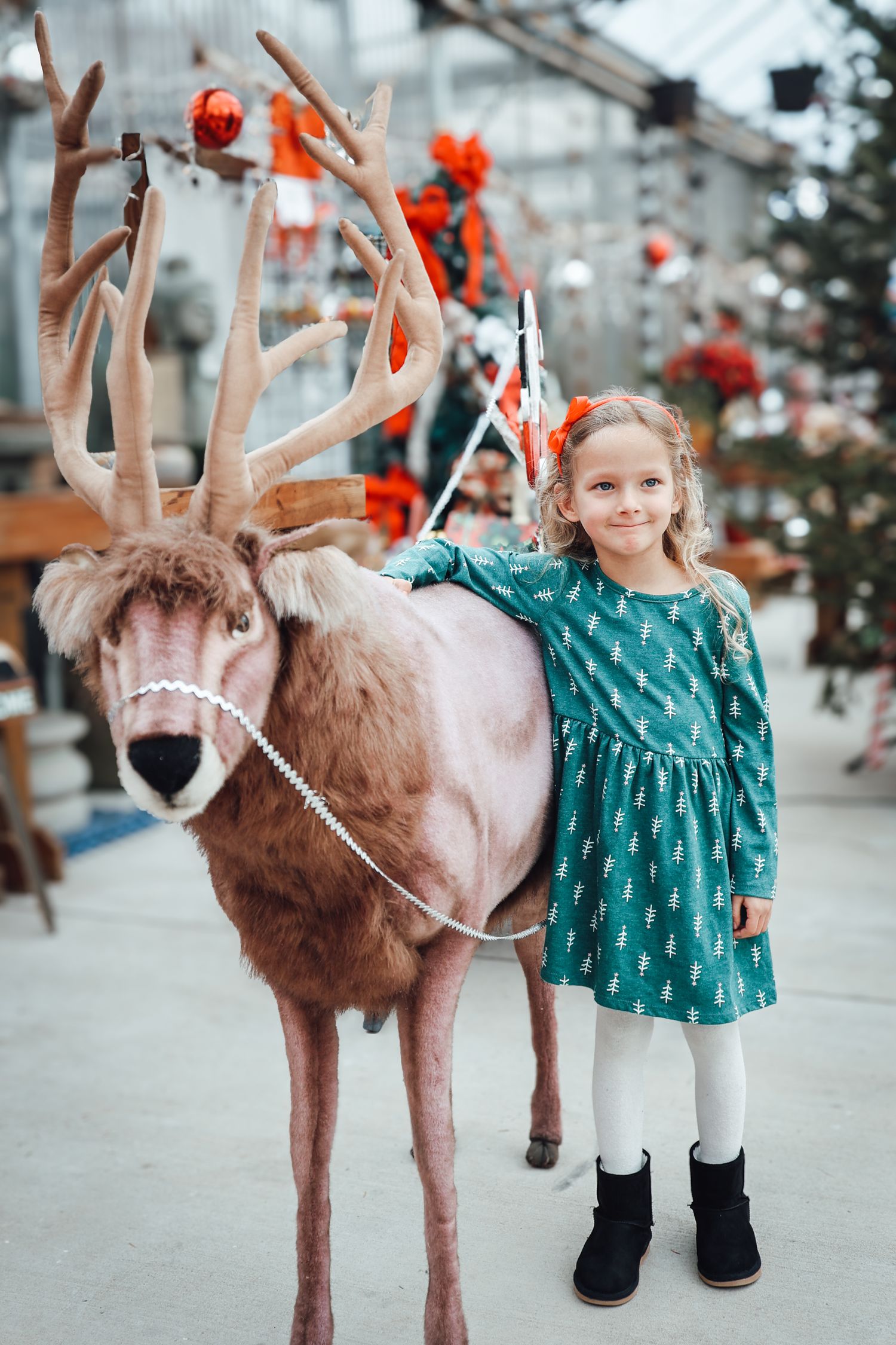 A child in a teal dress stands next to a decorative reindeer display with red ornaments and festive decor in the background.
