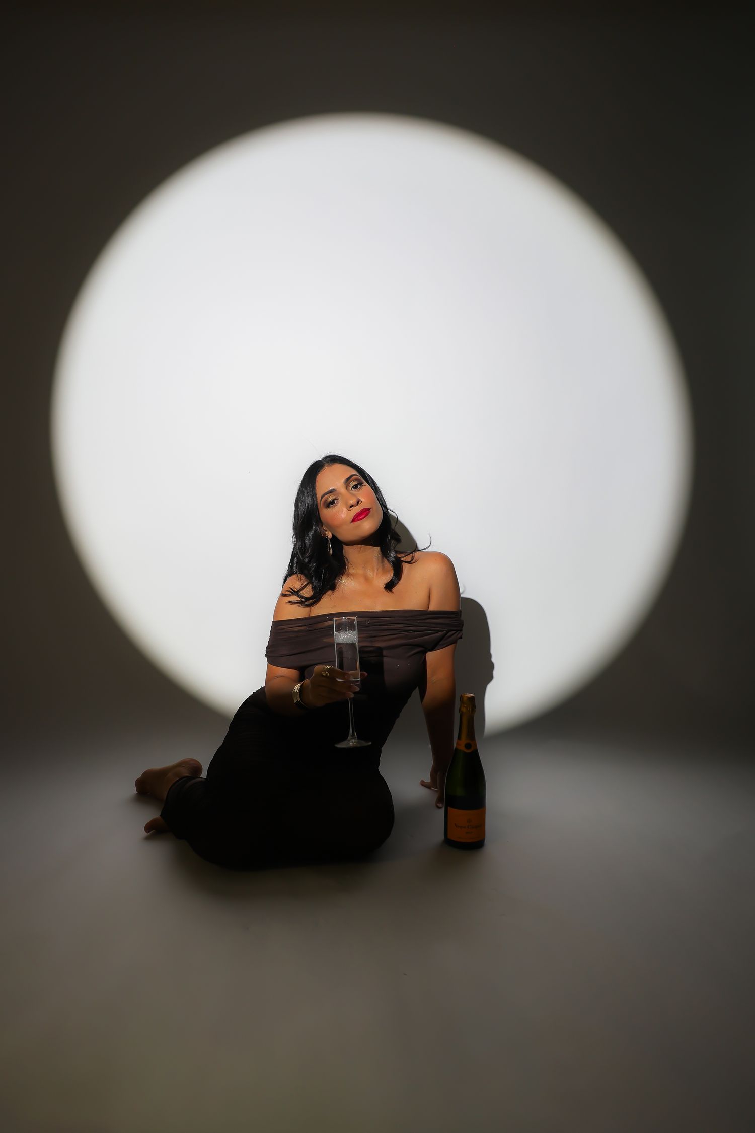 A figure in black clothing reclines beneath a dramatic spotlight on a dark stage in an artistic portrait series.
