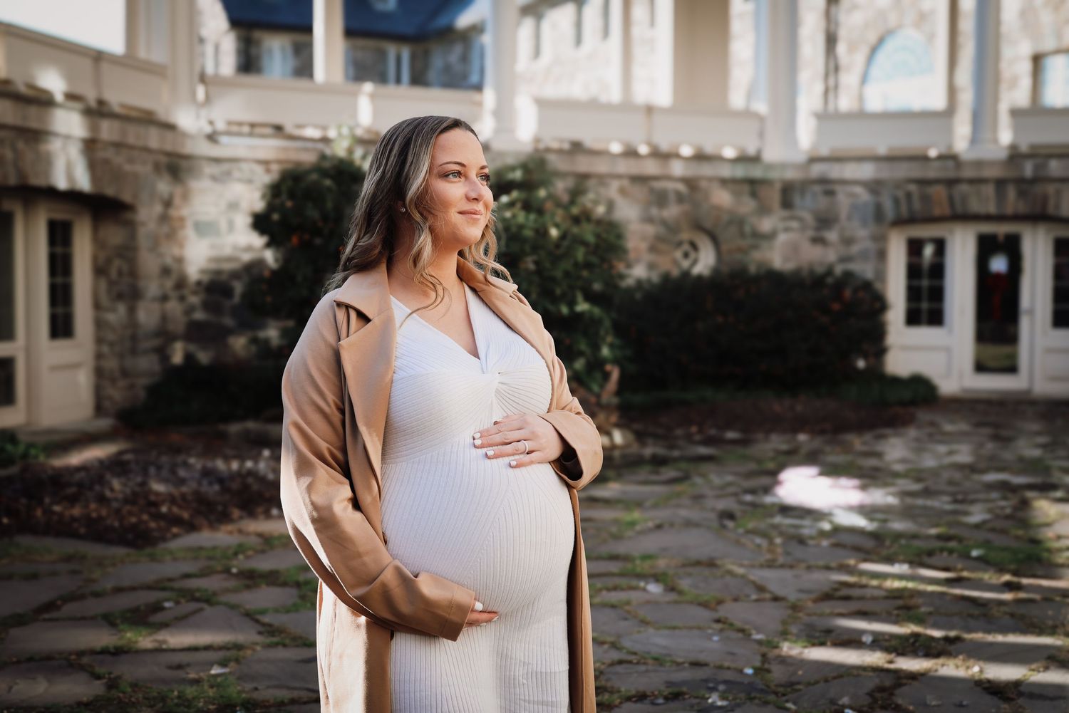 A pregnant woman in a white dress stands against a stone wall background in natural lighting.