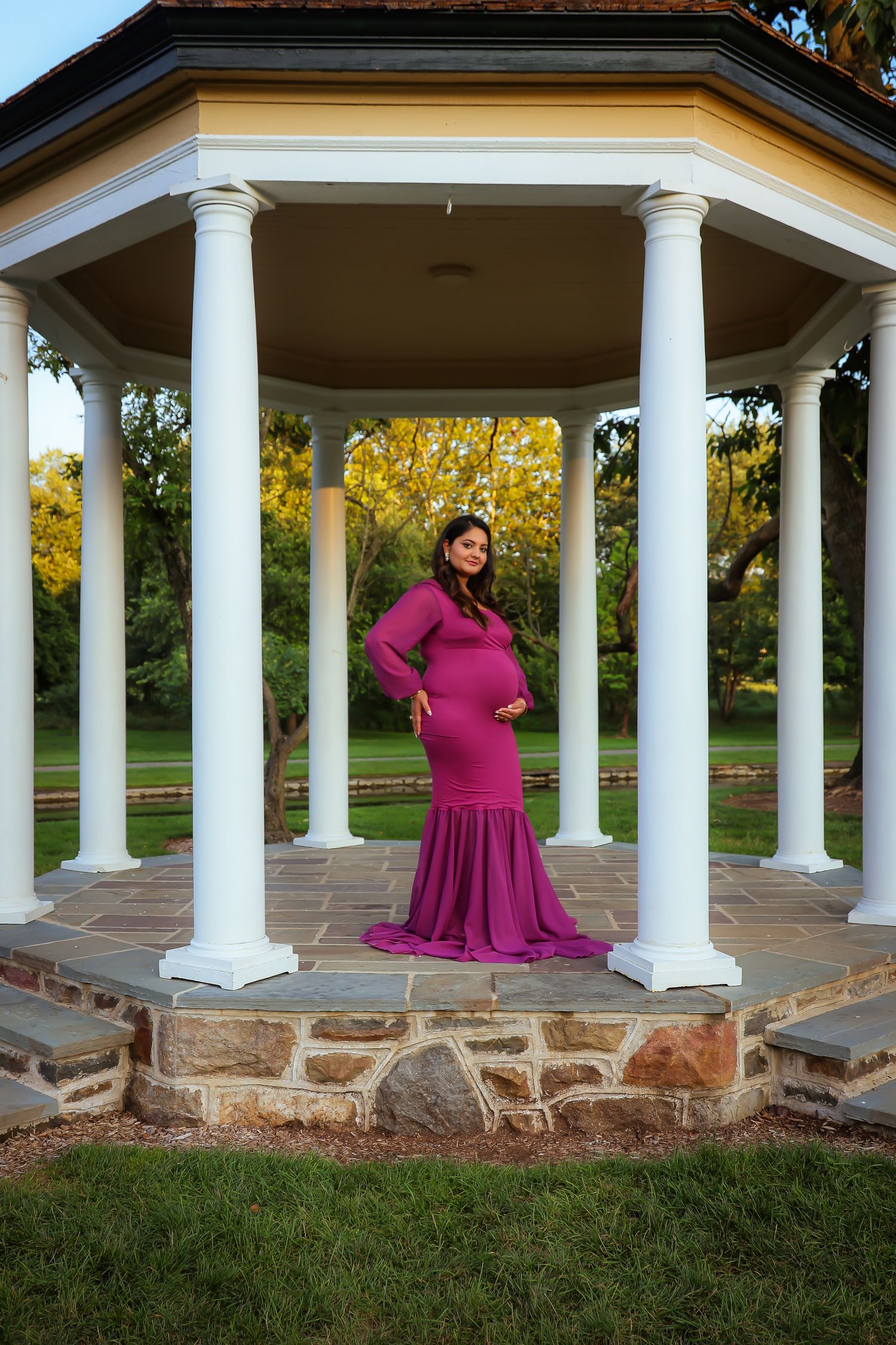 A figure in a flowing magenta gown poses elegantly in a white columned gazebo at sunset.
