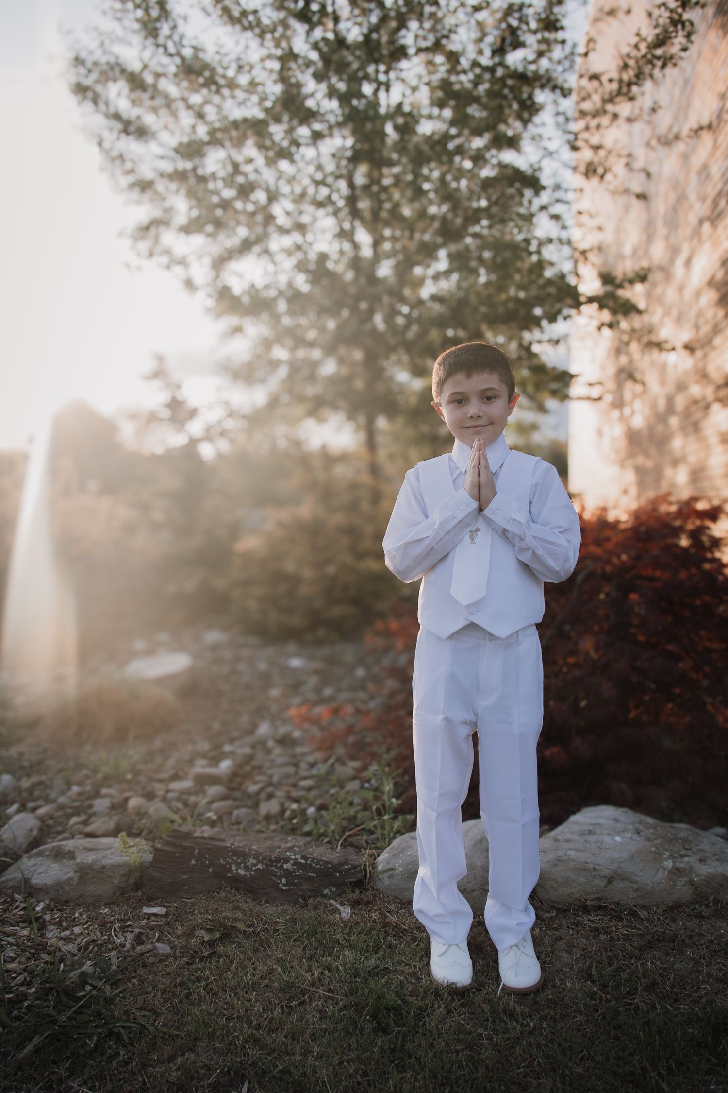 Young ring bearer in white suit stands on garden path during sunset wedding photoshoot.