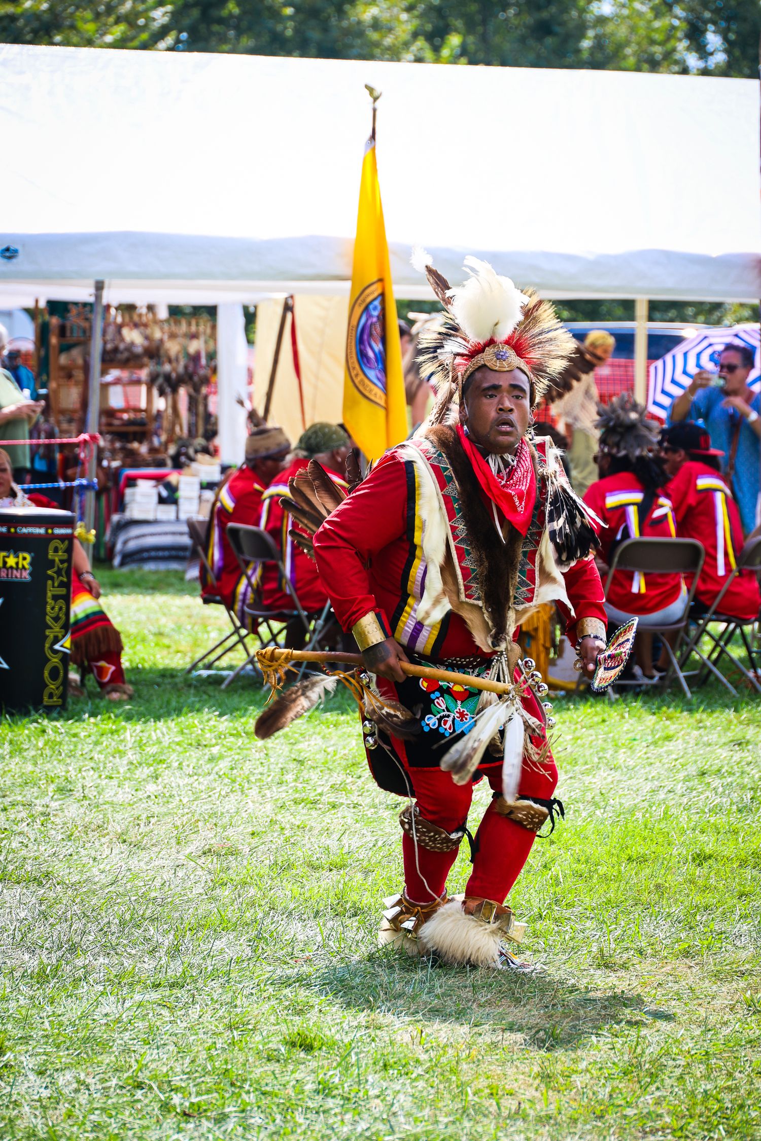 Native American dancer in traditional red regalia performs at outdoor cultural festival with tribal flag in background.