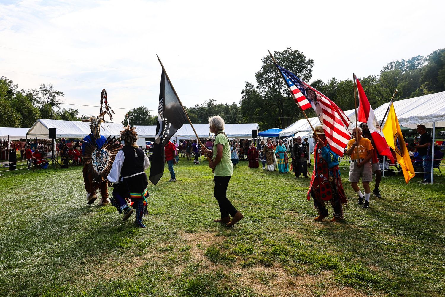 Native American dancers in traditional regalia perform at an outdoor powwow with flags and spectators visible.