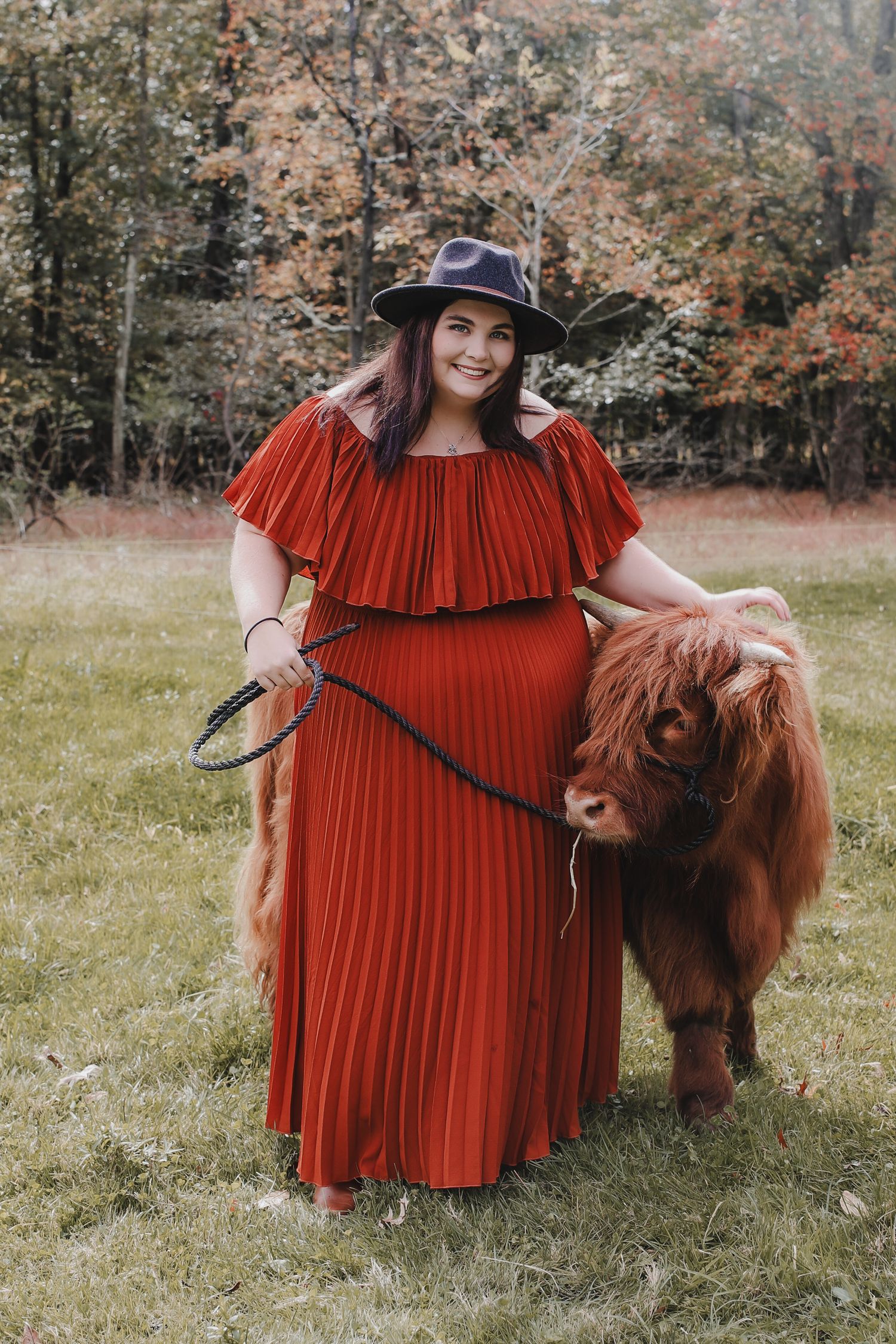 A person in a flowing red dress poses with a miniature pony in an autumn field setting.
