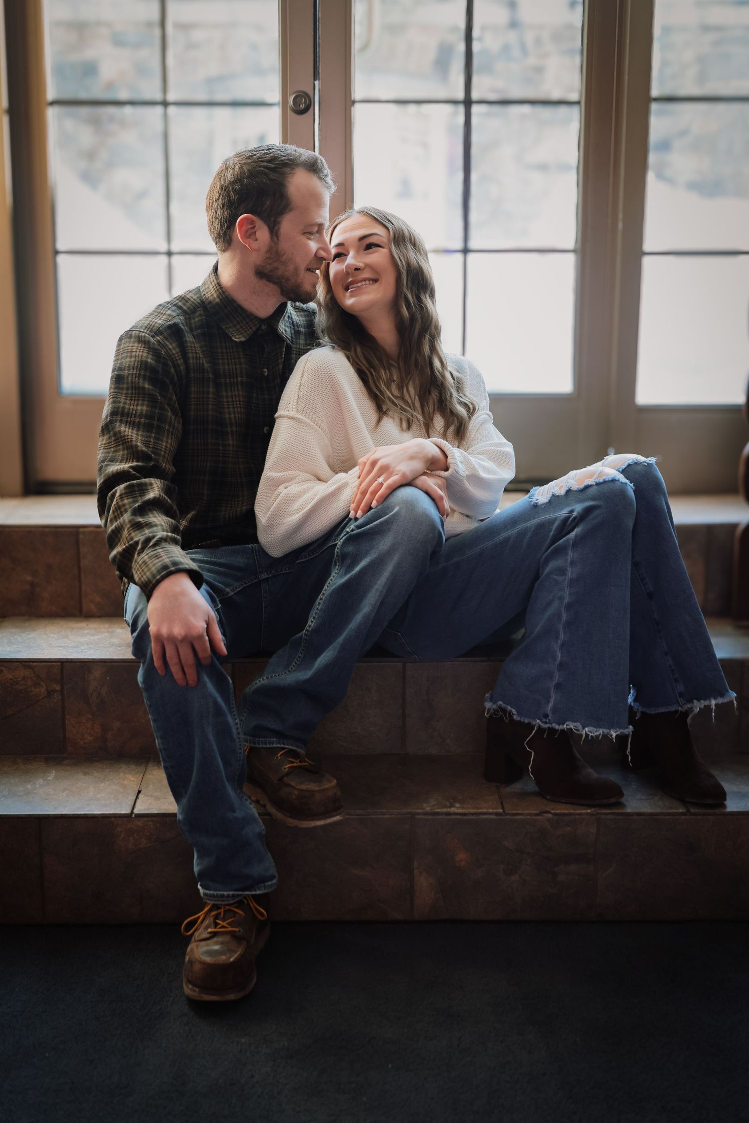 A couple sits intimately by a window ledge in casual clothing, creating a cozy and romantic atmosphere.