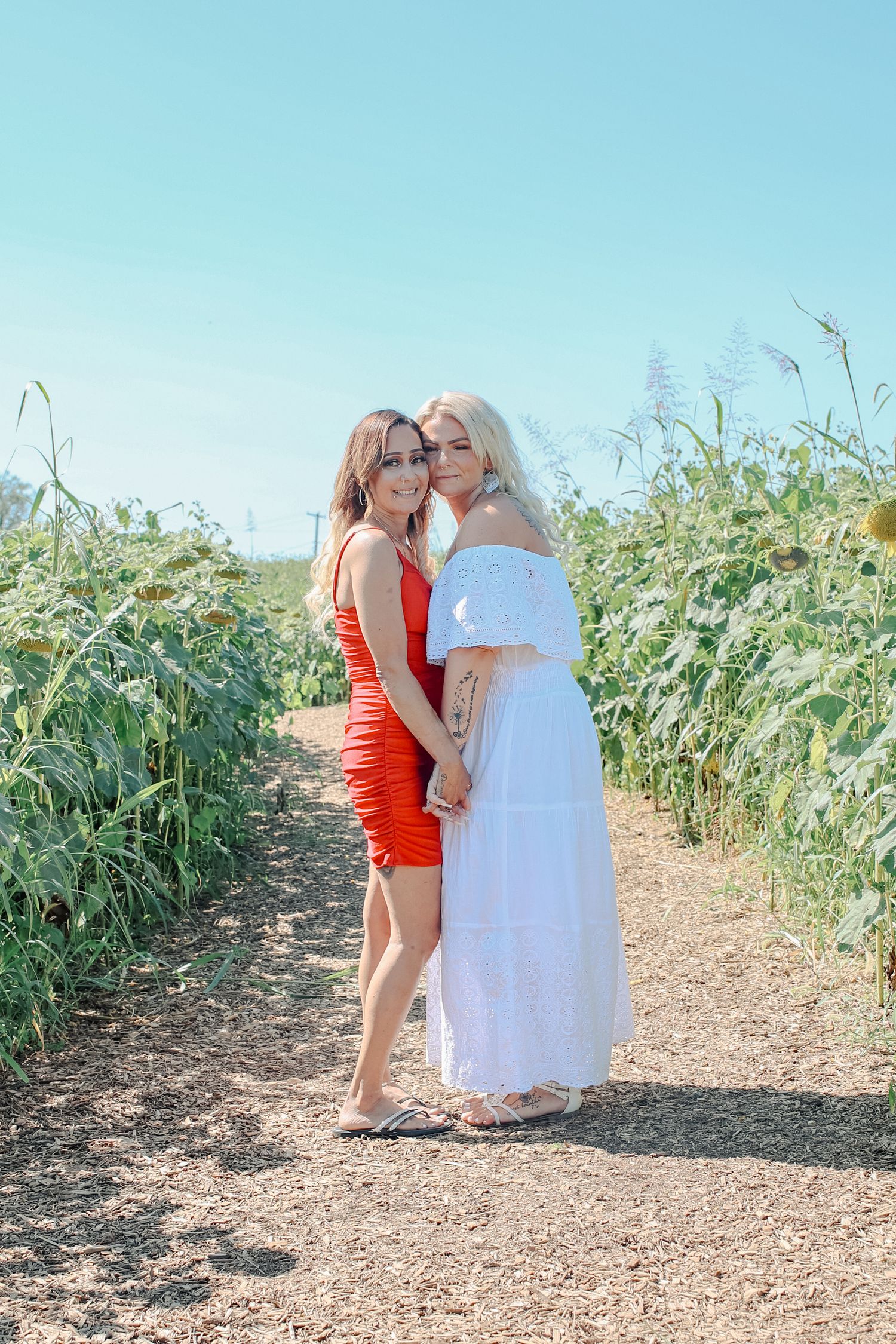 Two people in contrasting red and white dresses pose together on a dirt path surrounded by tall green plants on a sunny day.