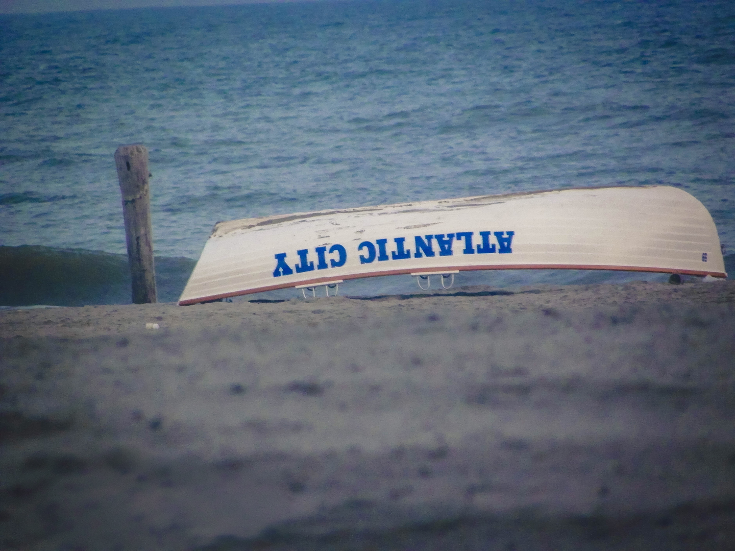 An Atlantic City lifeguard rowboat resting upside down on a sandy beach with ocean waves in the background.