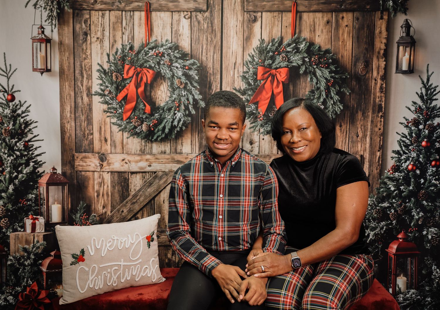 A couple sits on a red bench in matching plaid outfits against a rustic holiday backdrop with wreaths and lanterns.