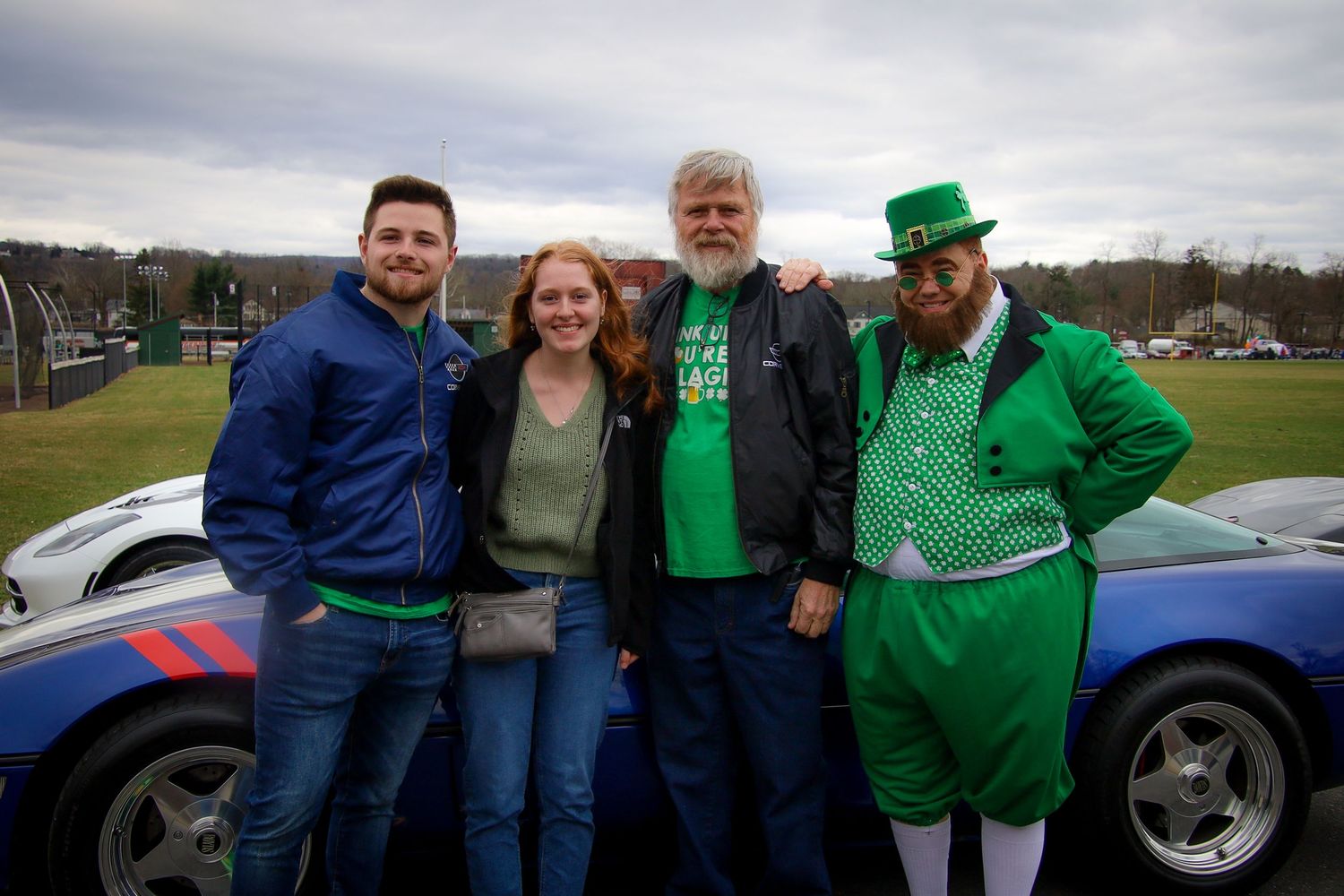 People wearing festive green attire gather around cars at an outdoor St. Patrick's Day event.