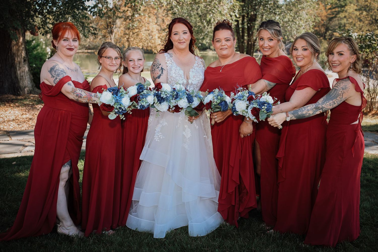 A wedding party poses outdoors wearing burgundy bridesmaid dresses and a white wedding gown with floral accents.