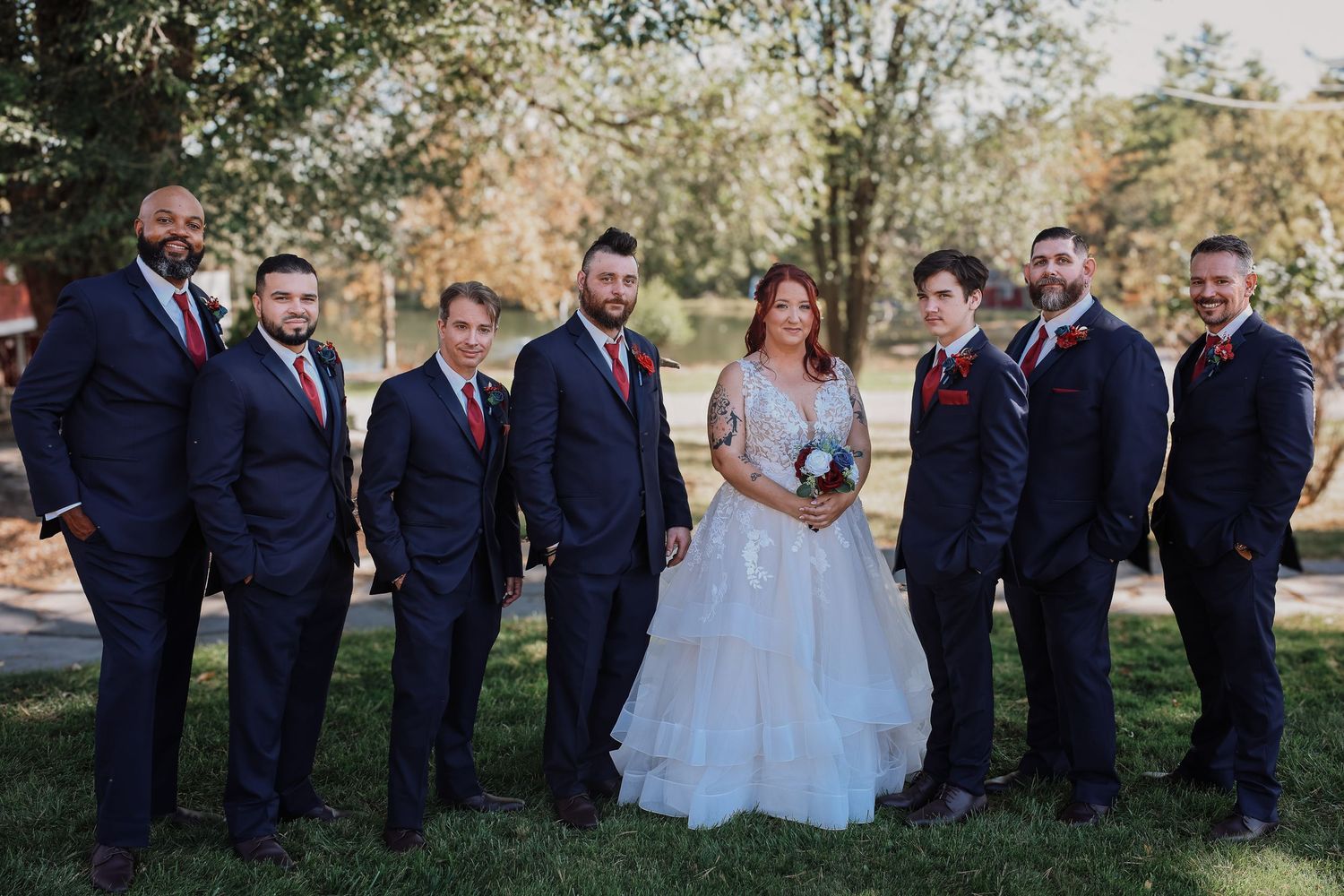 Wedding party in navy suits and white dresses pose together on a grassy lawn during an outdoor celebration.