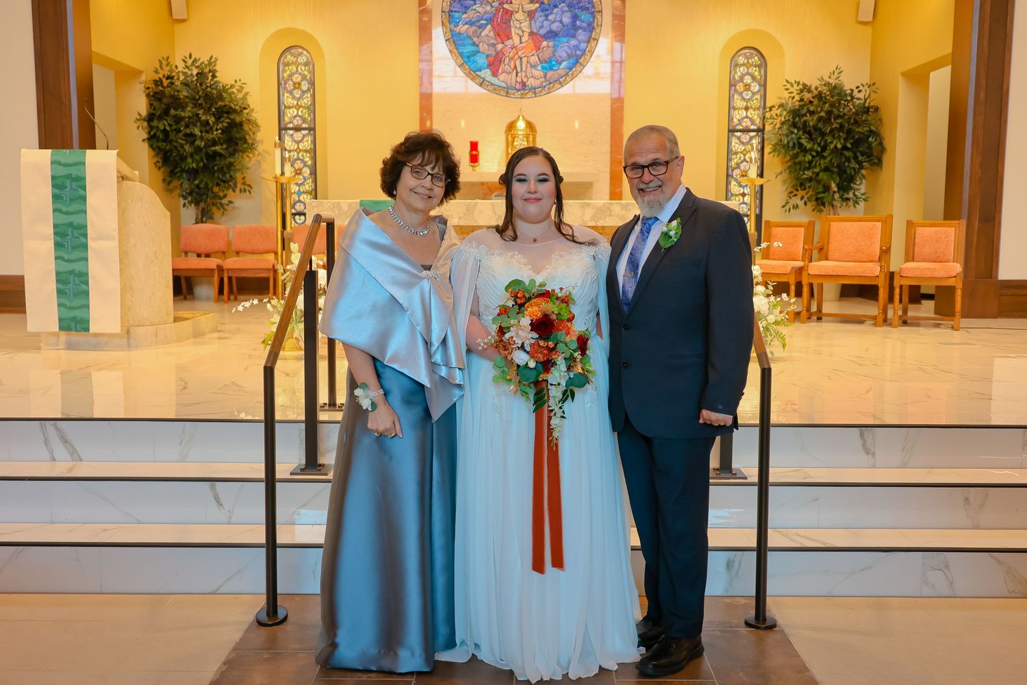 Three people in formal attire pose together at the altar of a church with stained glass and decorative plants.