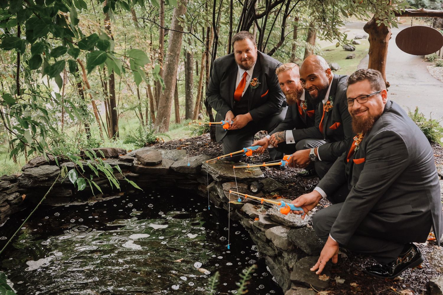 Three groomsmen in dark suits and orange ties gather around a decorative pond in a garden setting.