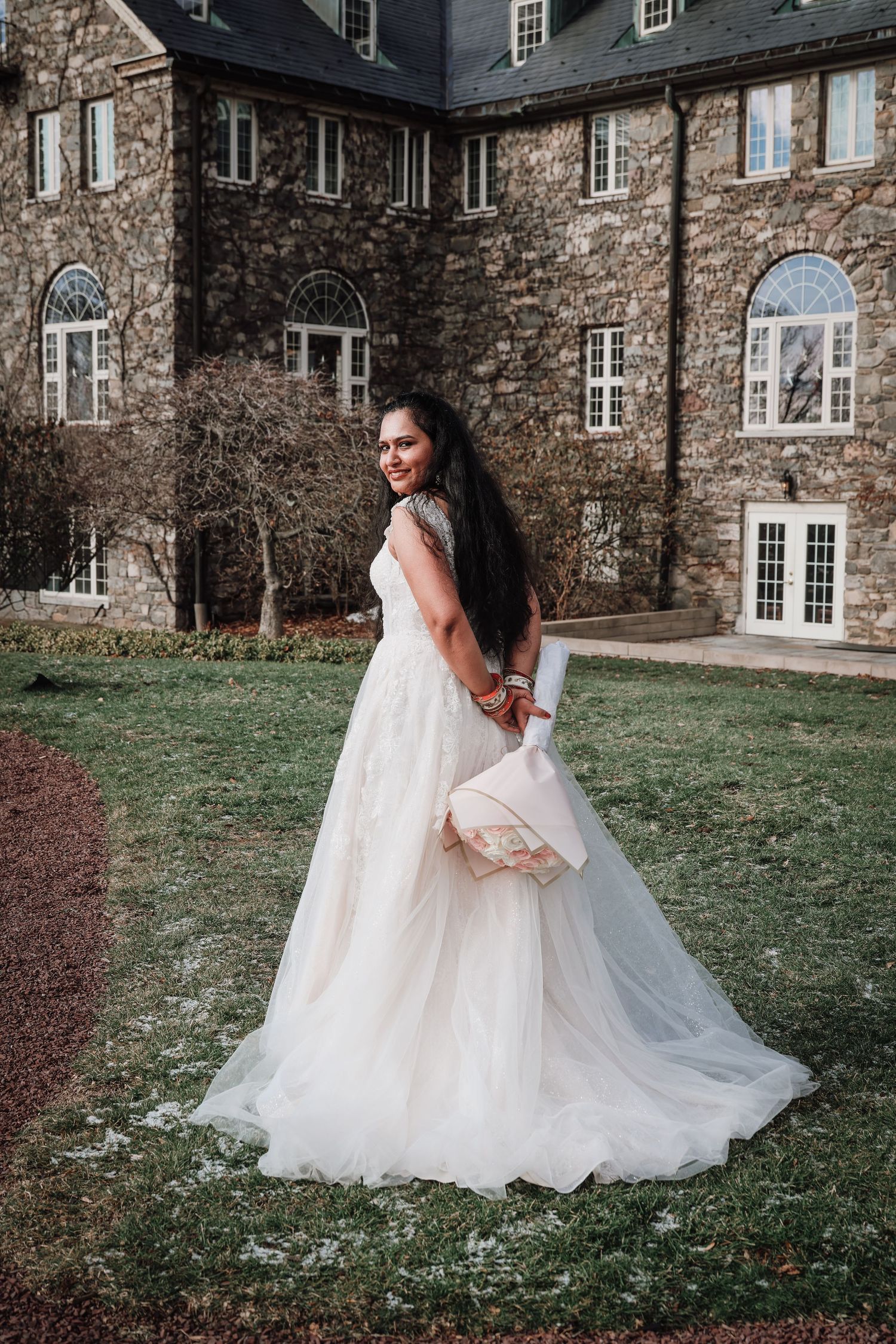 A bride in a flowing white wedding gown twirls gracefully on the lawn in front of a historic stone mansion.