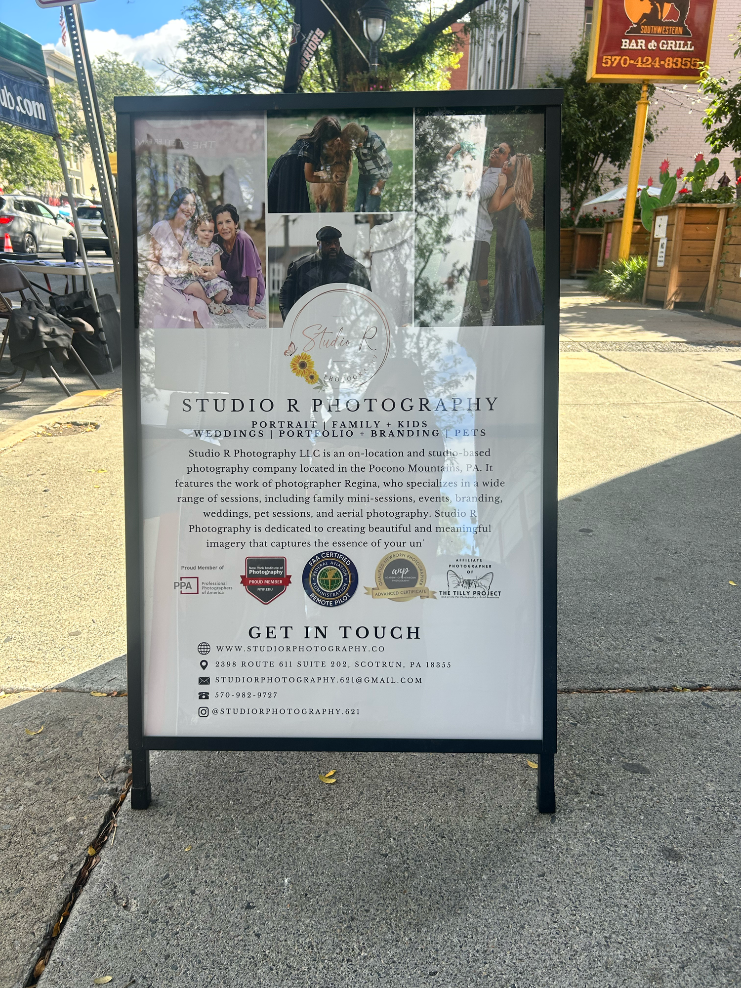 A sidewalk A-frame sign displays studio photography services and contact information with sample photos and professional logos.
