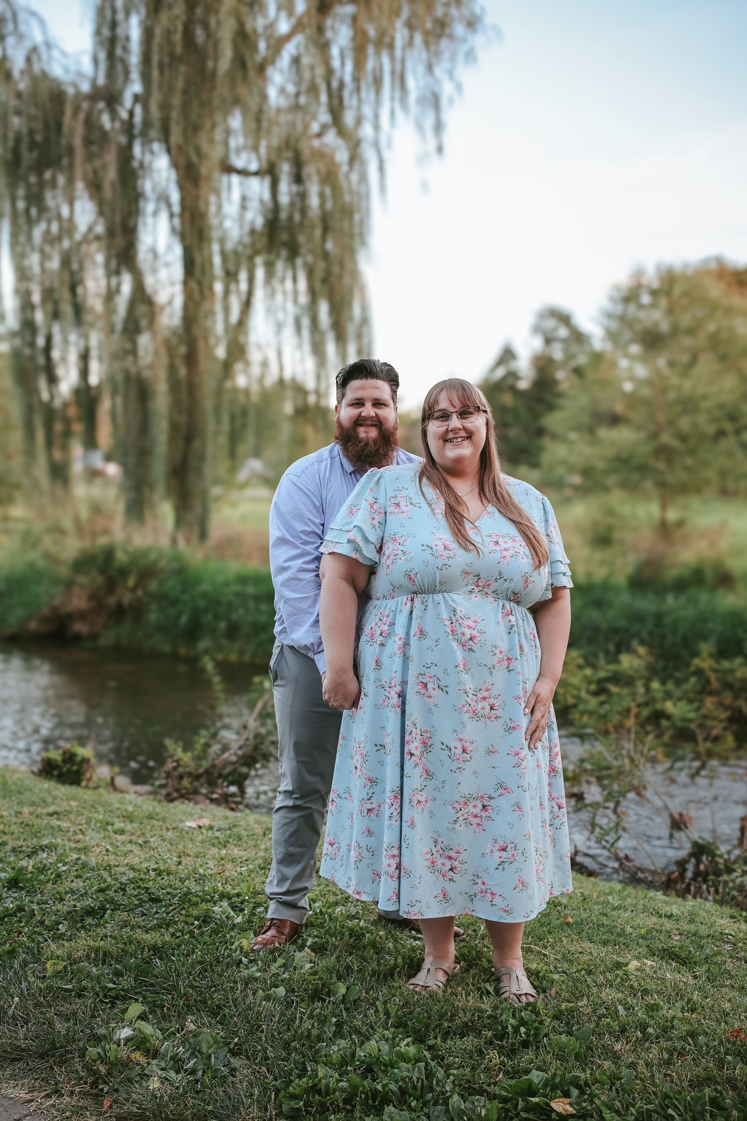A couple poses together near a willow tree and pond during an outdoor photo session in a romantic setting.