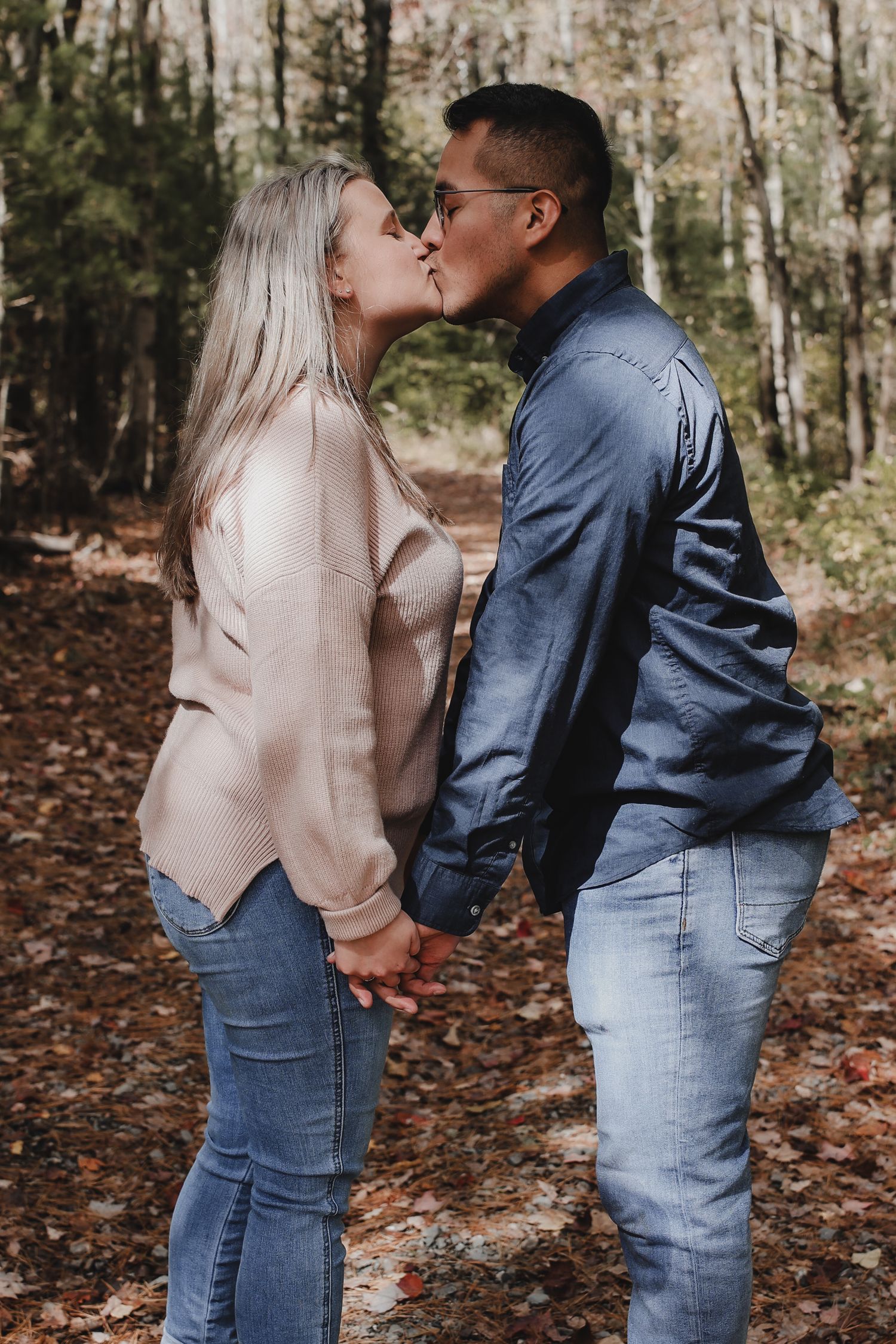 A couple in denim and sweaters share tender moments in a wooded autumn setting with fallen leaves on the ground.