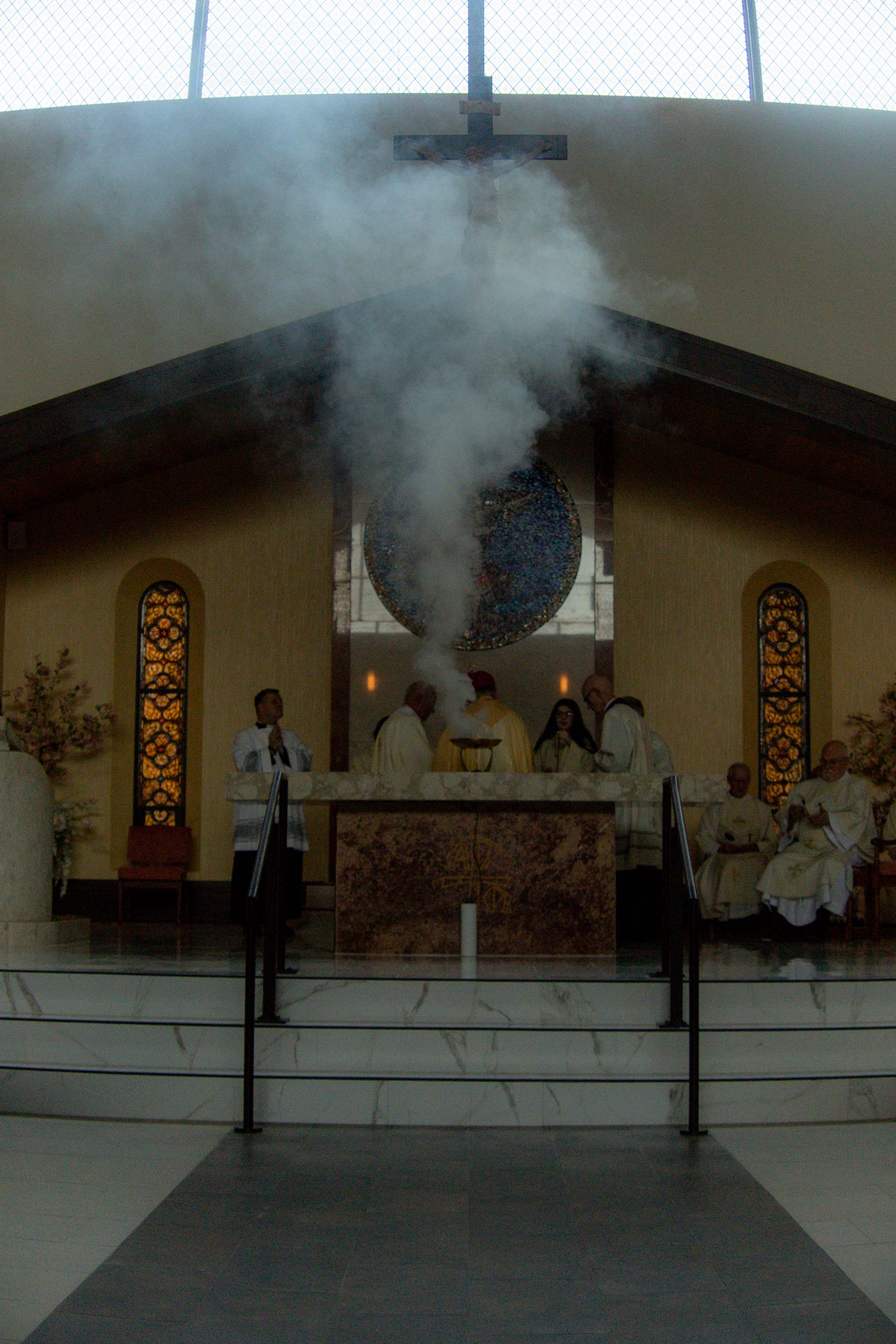 A sequence of dark silhouettes move through a smoky indoor space with decorative stained glass windows visible in the background.