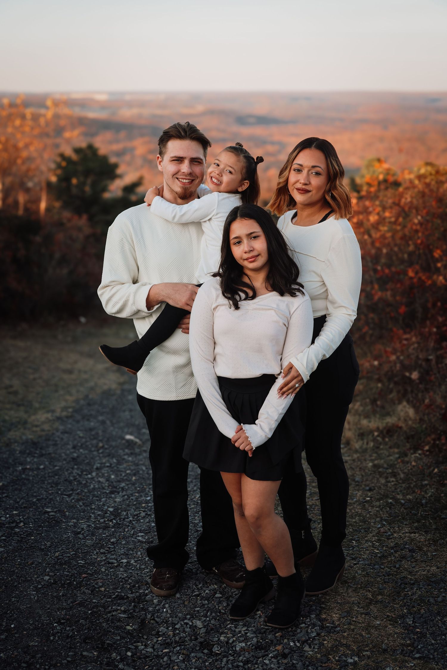 A family in matching white sweaters and black pants poses together outdoors during golden hour autumn sunset.