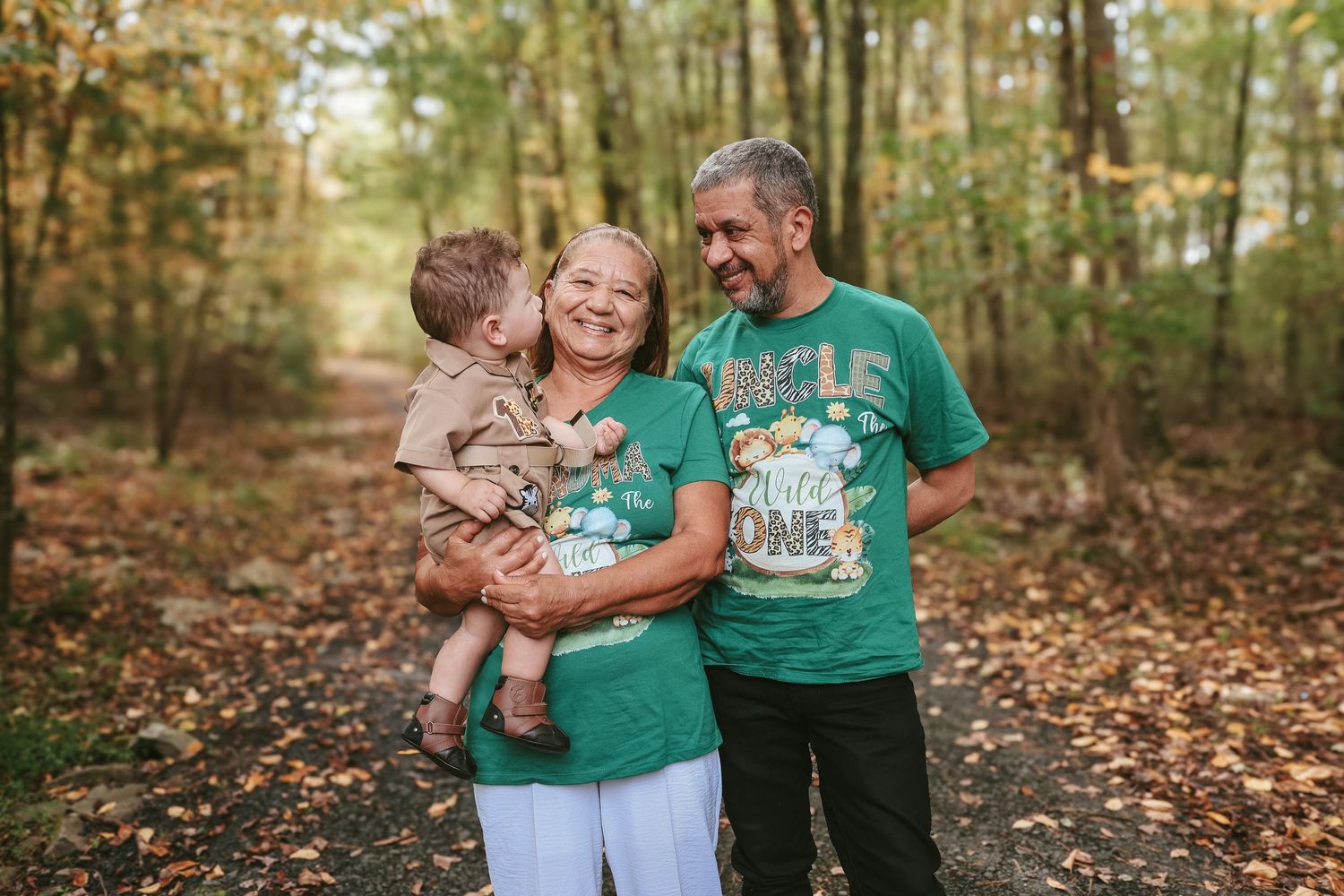 Family wearing coordinated teal and white outfits embraces while standing in autumn forest with fallen leaves.