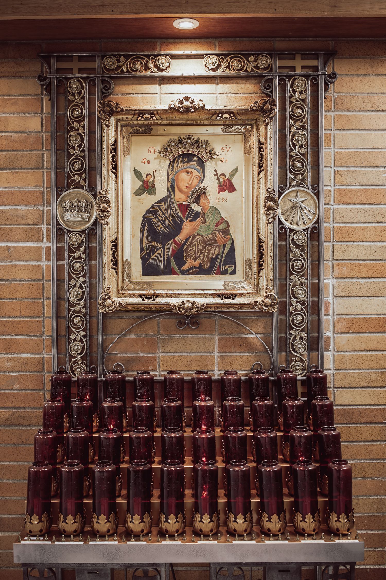 Religious icon of Mary and Jesus displayed in ornate gold frame above rows of red prayer candles in church alcove.