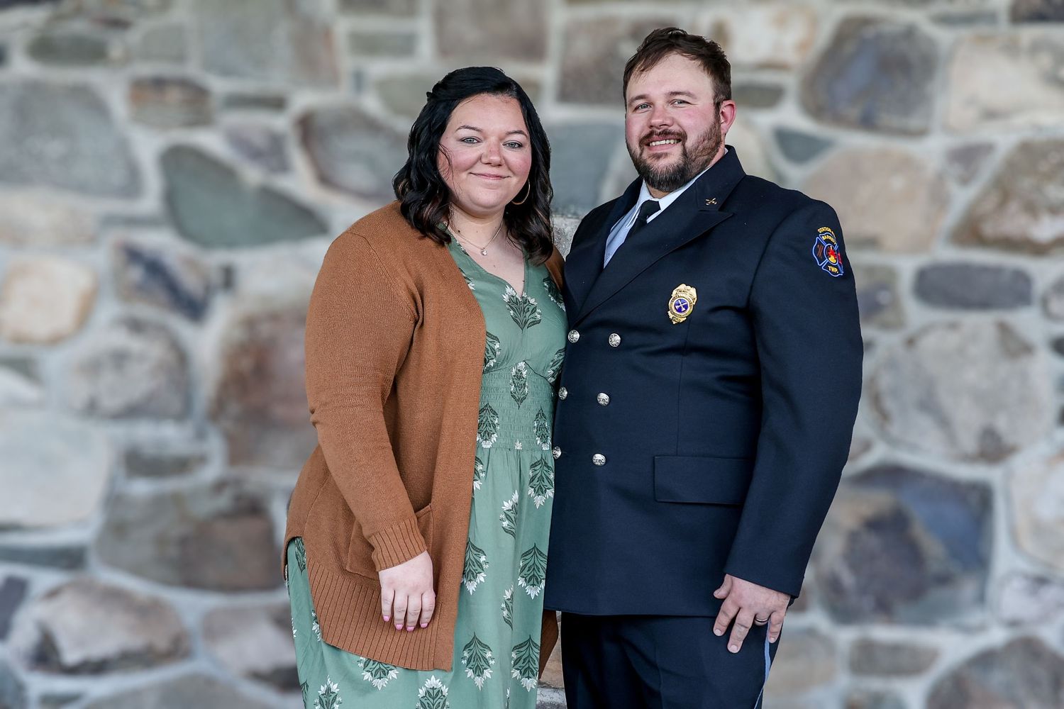 A couple poses together in front of a stone wall while wearing casual and formal attire.