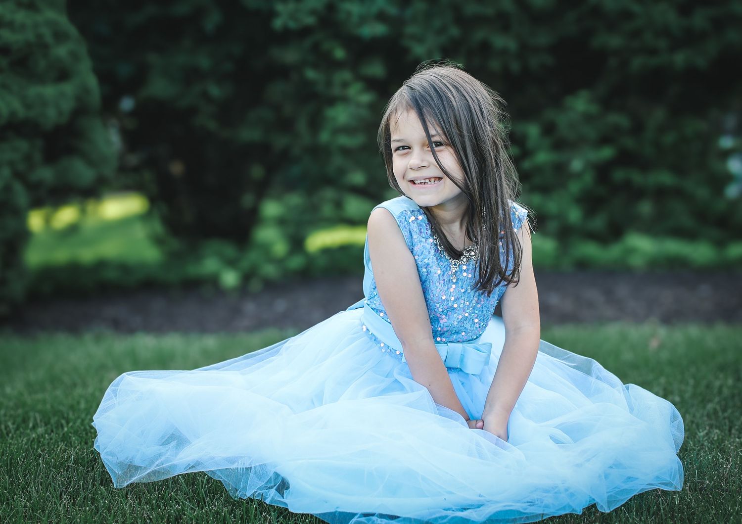 A person in a flowing light blue dress sits gracefully on green grass in a garden setting.