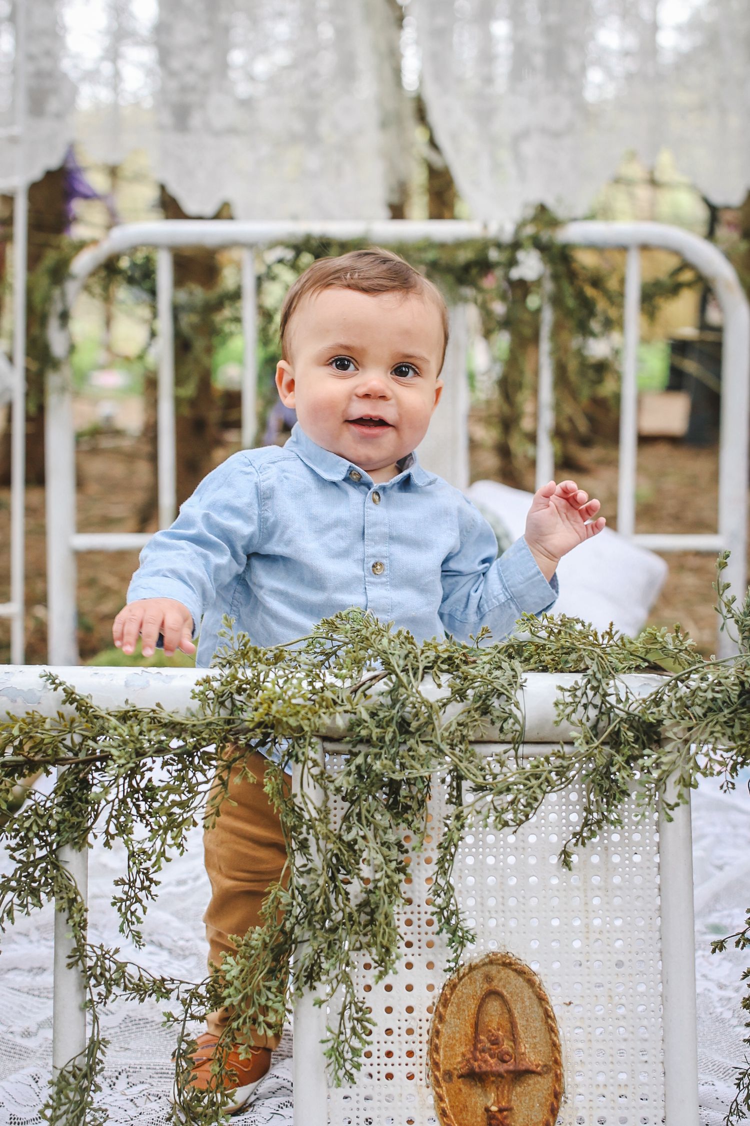 A baby in a light blue shirt and khaki pants sits on a white lattice bench decorated with greenery.