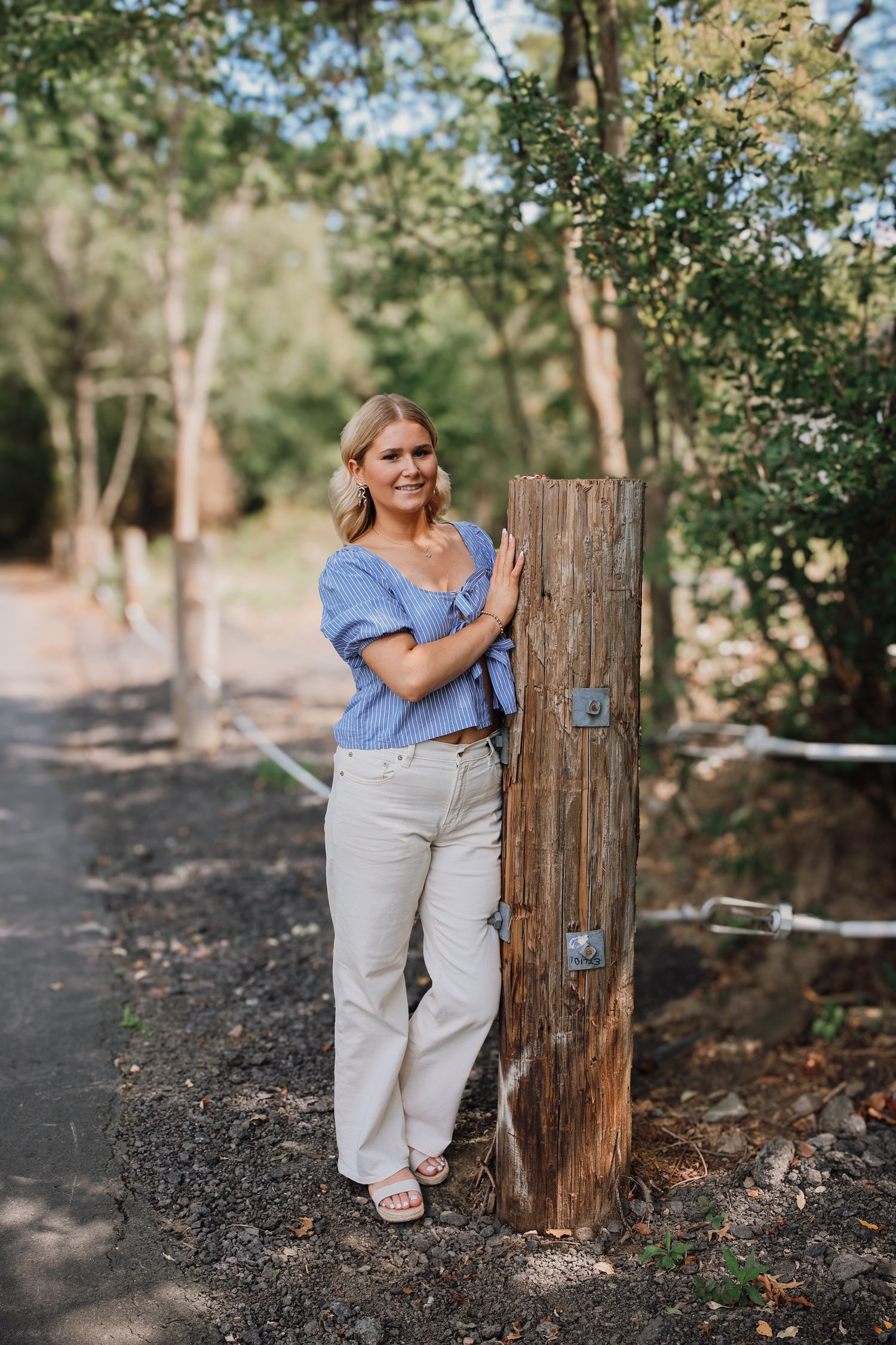 A person in casual attire poses against a rustic wooden post in an outdoor setting with dappled sunlight.