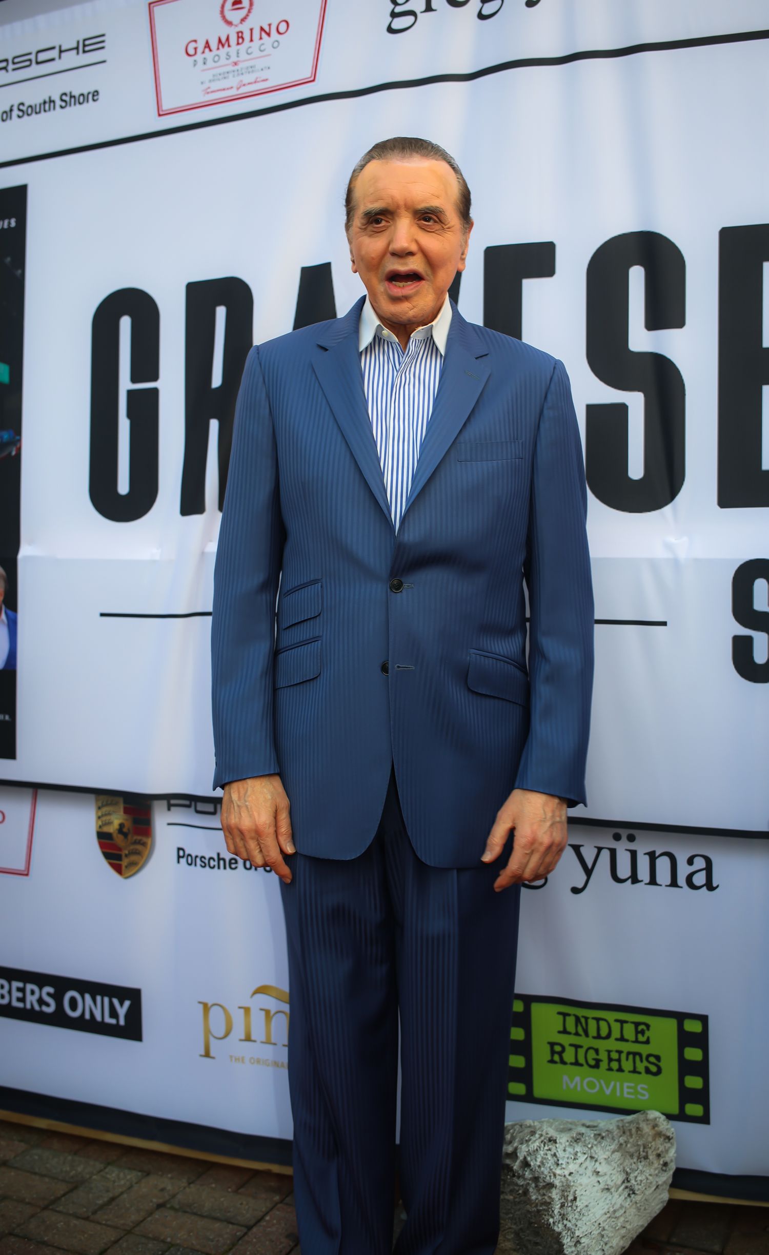 A person in a navy blue suit and striped dress shirt stands in front of a promotional backdrop at an event.