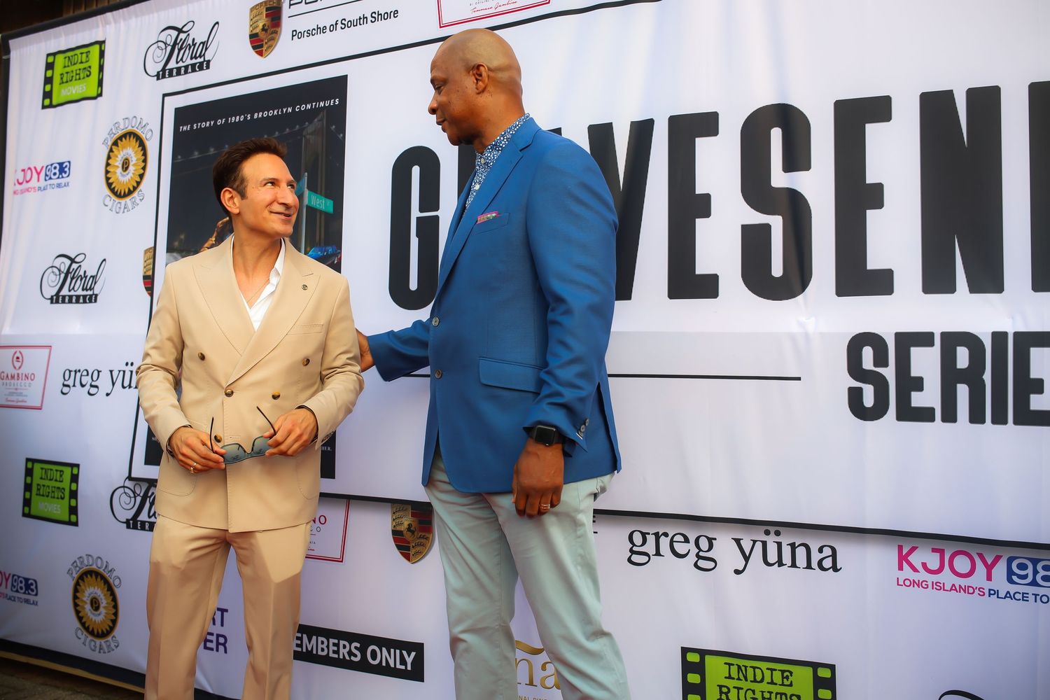 Two people in formal attire stand together at a promotional event with a large text display behind them.