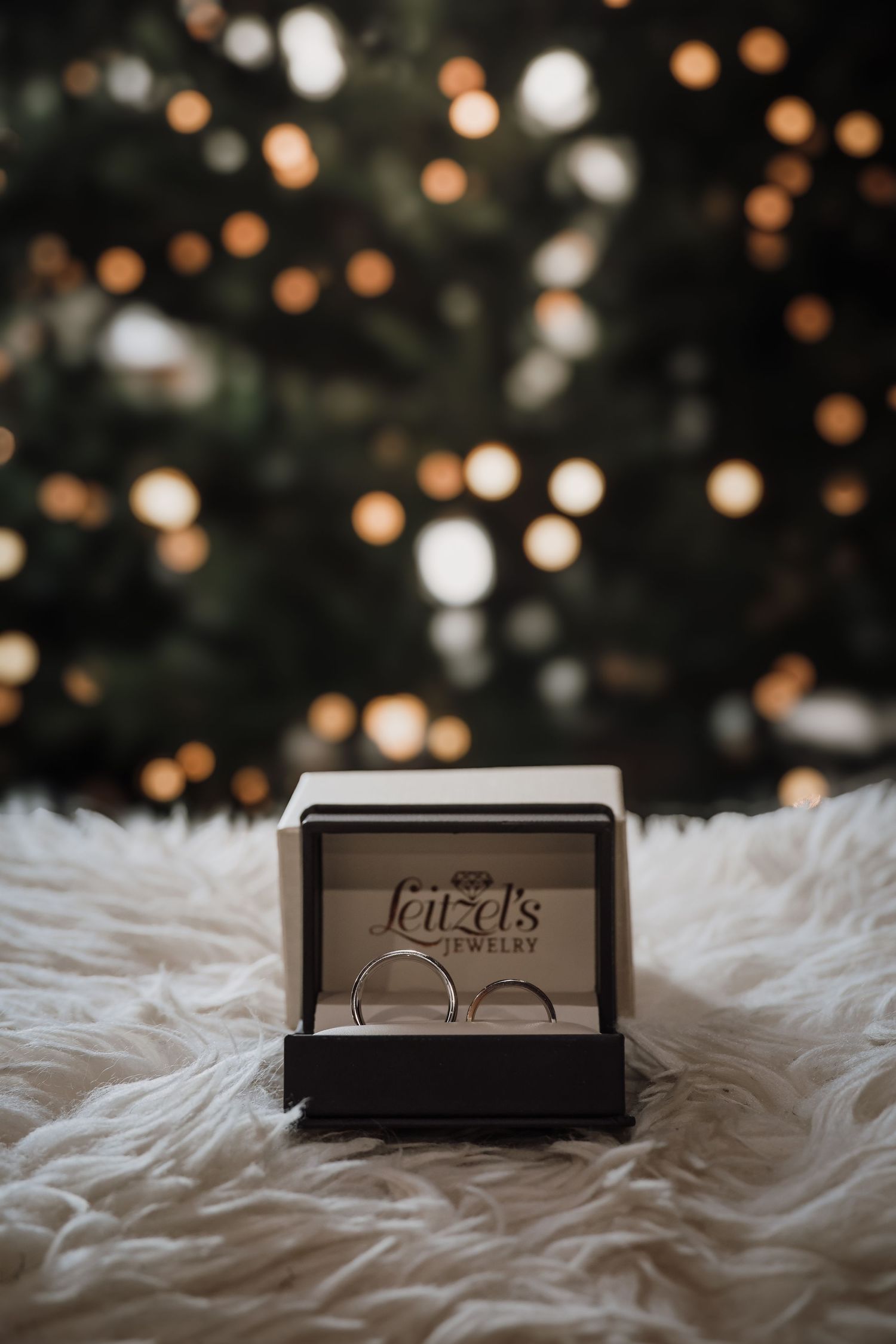 A minimalist wedding ring box sits on white fur with bokeh lights in the background creating a romantic atmosphere.
