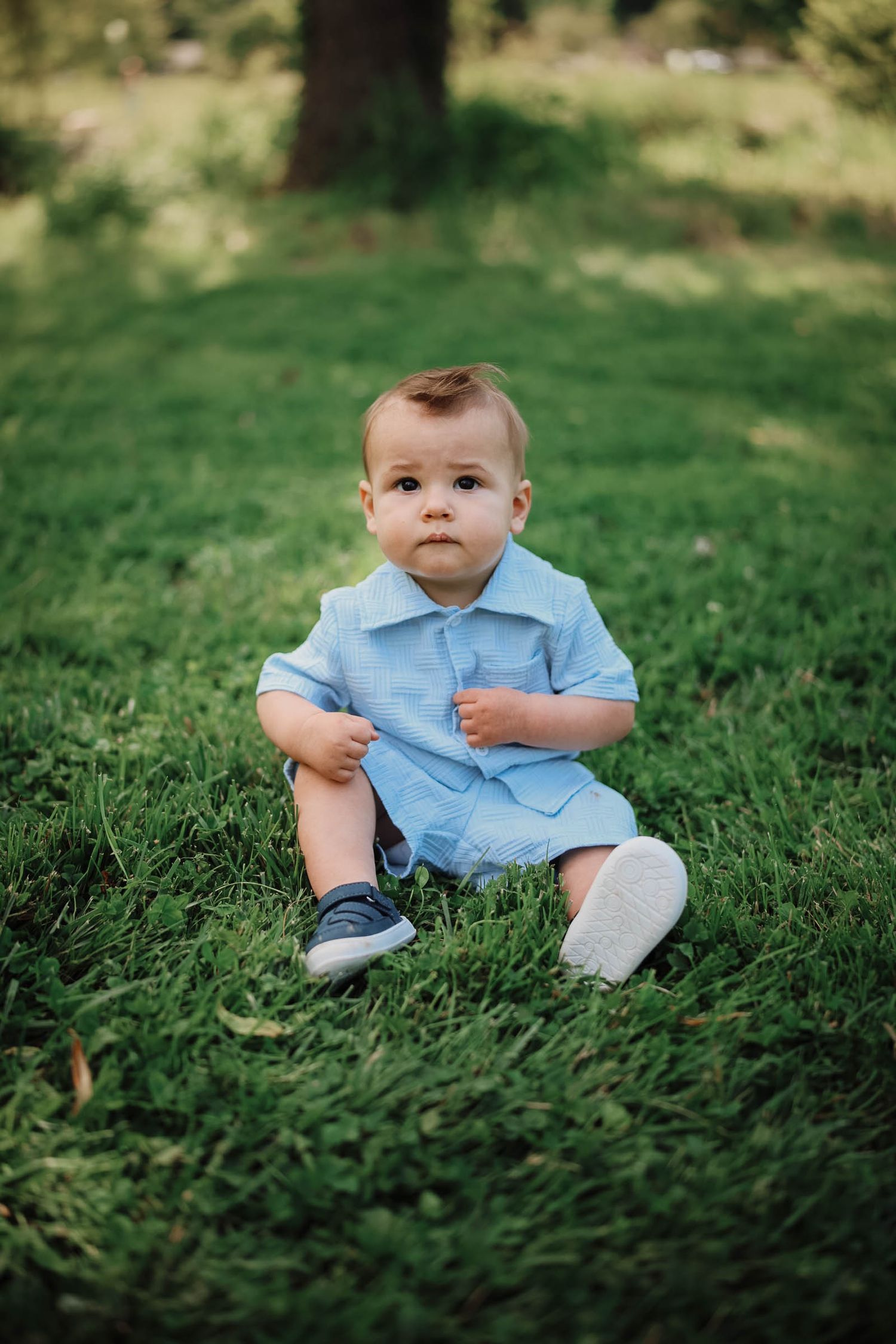 A cute baby boy in a blue romper sits on green grass in a sunny outdoor park setting.
