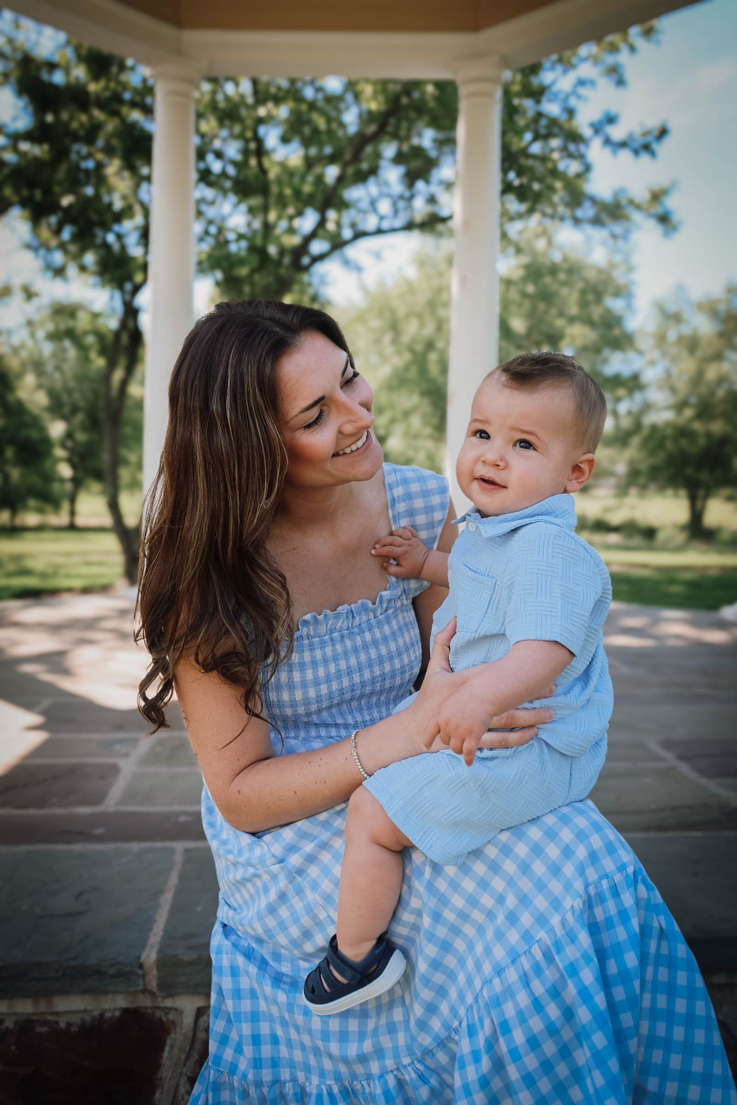 A smiling mother in a blue gingham dress holds her baby boy outdoors near a white gazebo.
