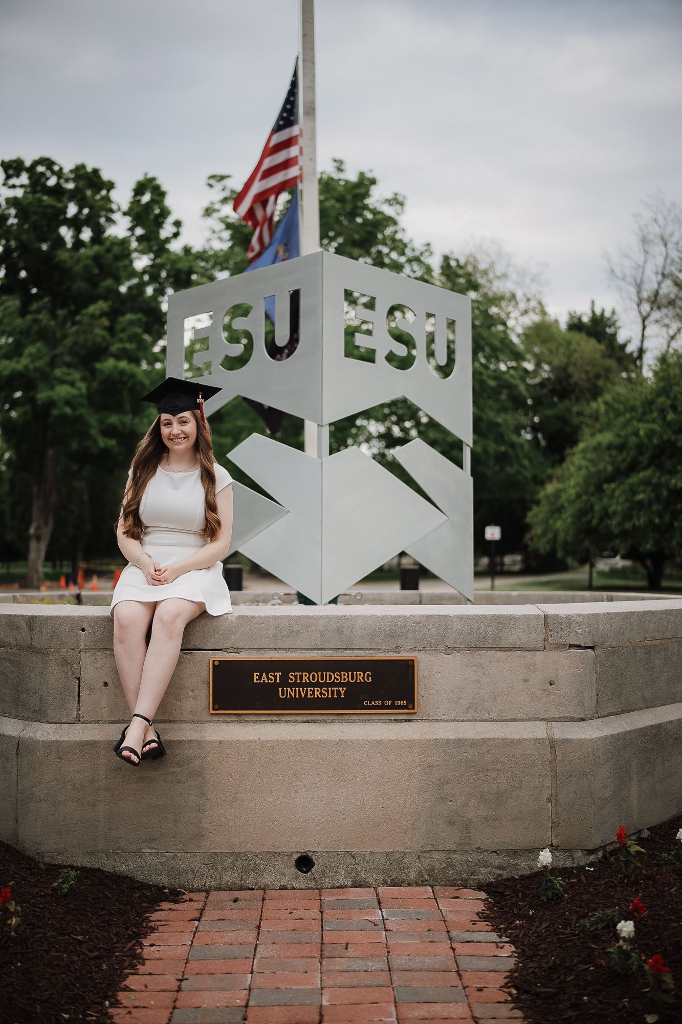 A smiling graduate in cap and white dress sits at East Stroudsburg University sign with American flag backdrop.