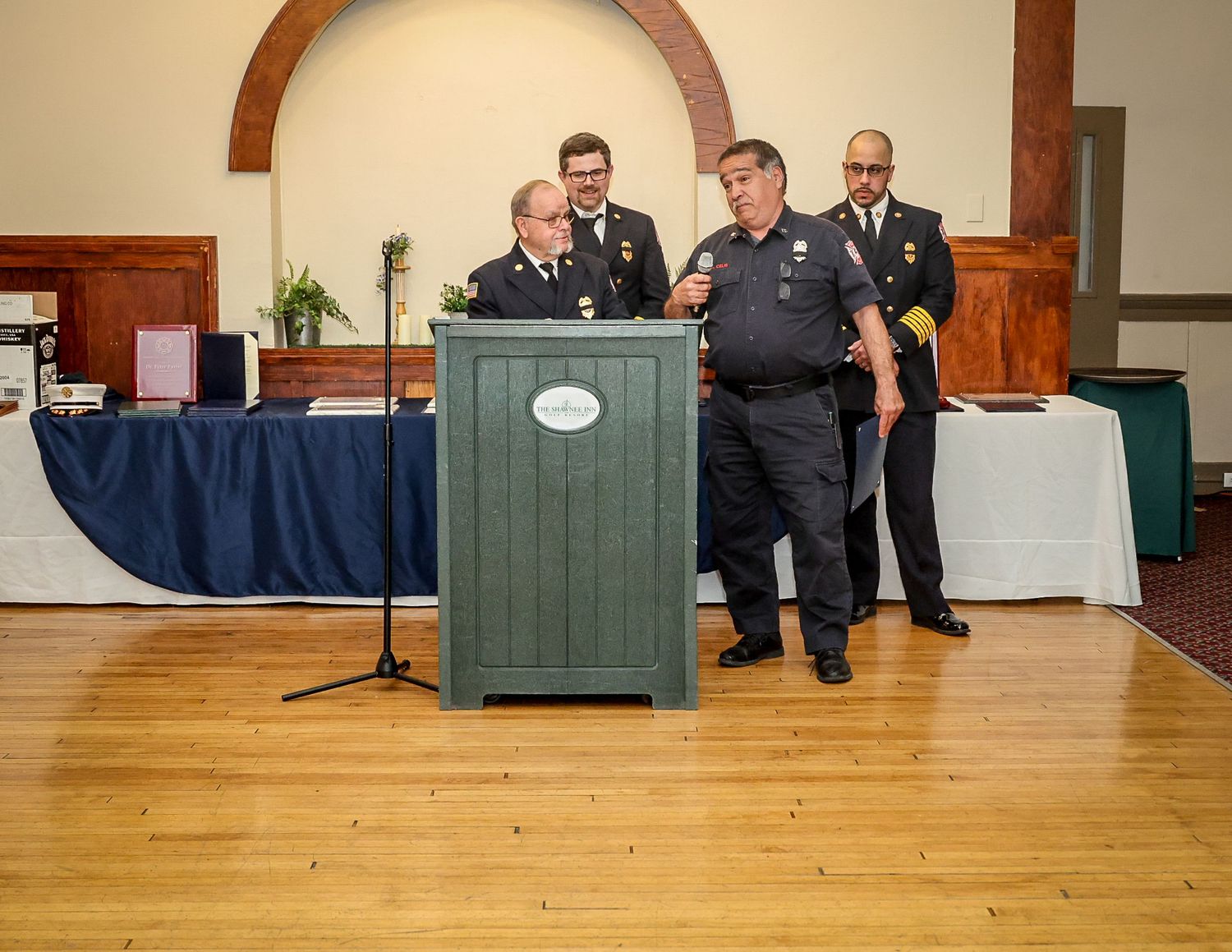 Officials in uniforms stand at a green podium during a formal ceremony in a wood-floored hall.