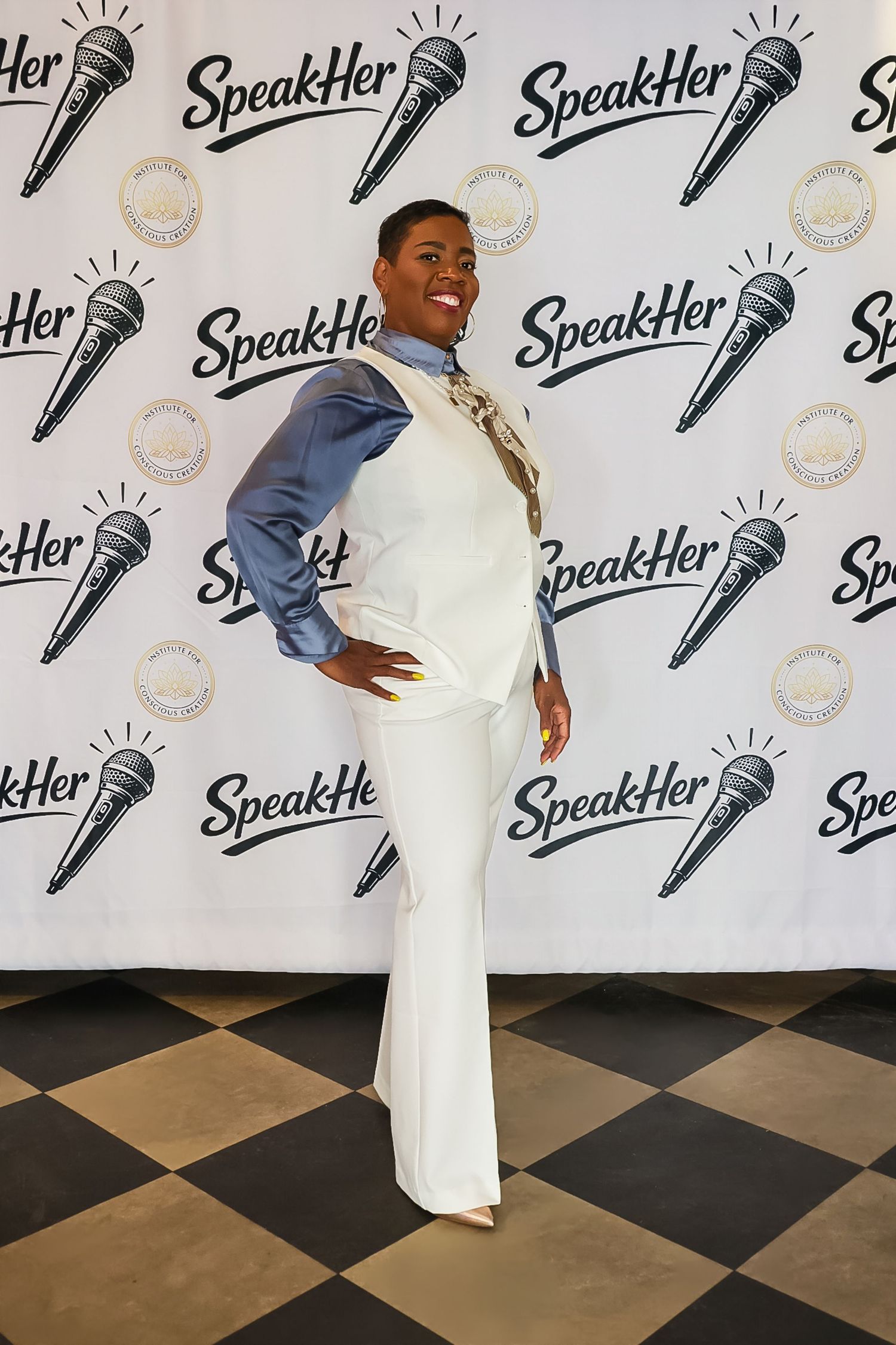Woman in white outfit poses at SpeakHer event backdrop, wearing blue denim jacket and gold scarf accessories.