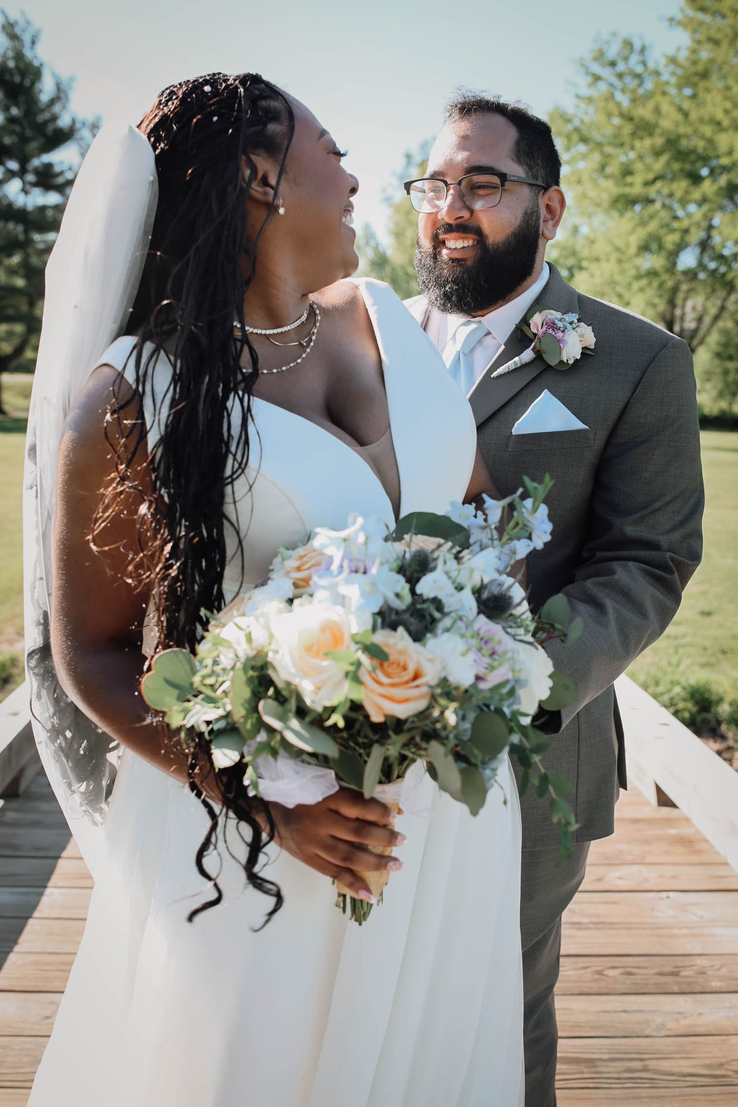 A bride in a white gown holding a floral bouquet smiles with a bearded groom in a gray suit on a wooden bridge.