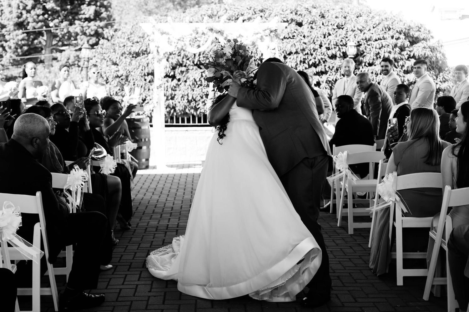 Black and white photo of bride and groom sharing first kiss at outdoor wedding ceremony with guests seated in white chairs.