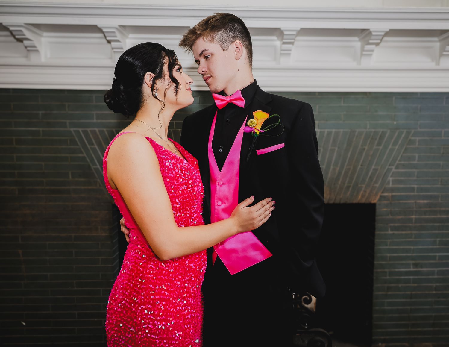 Couple in matching hot pink prom attire, sequined dress and tuxedo with bow tie, sharing a romantic moment.