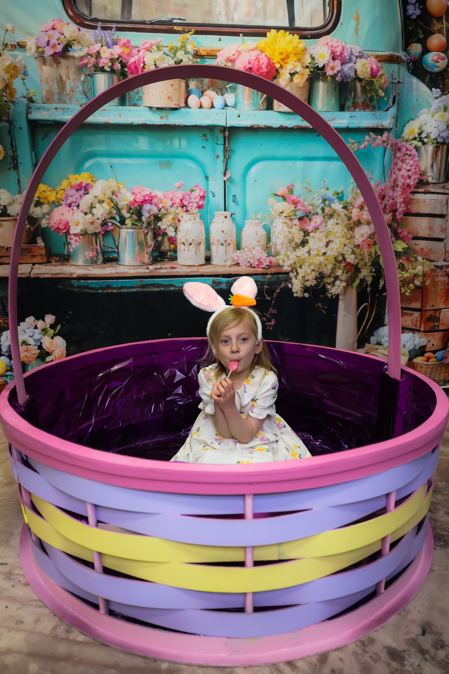 Girl in bunny ears sits inside a giant colorful Easter basket with floral backdrop and Easter eggs.