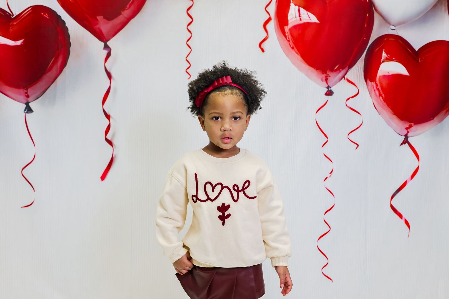 Young girl in a cream love sweatshirt poses with red heart balloons for Valentine's Day photo.