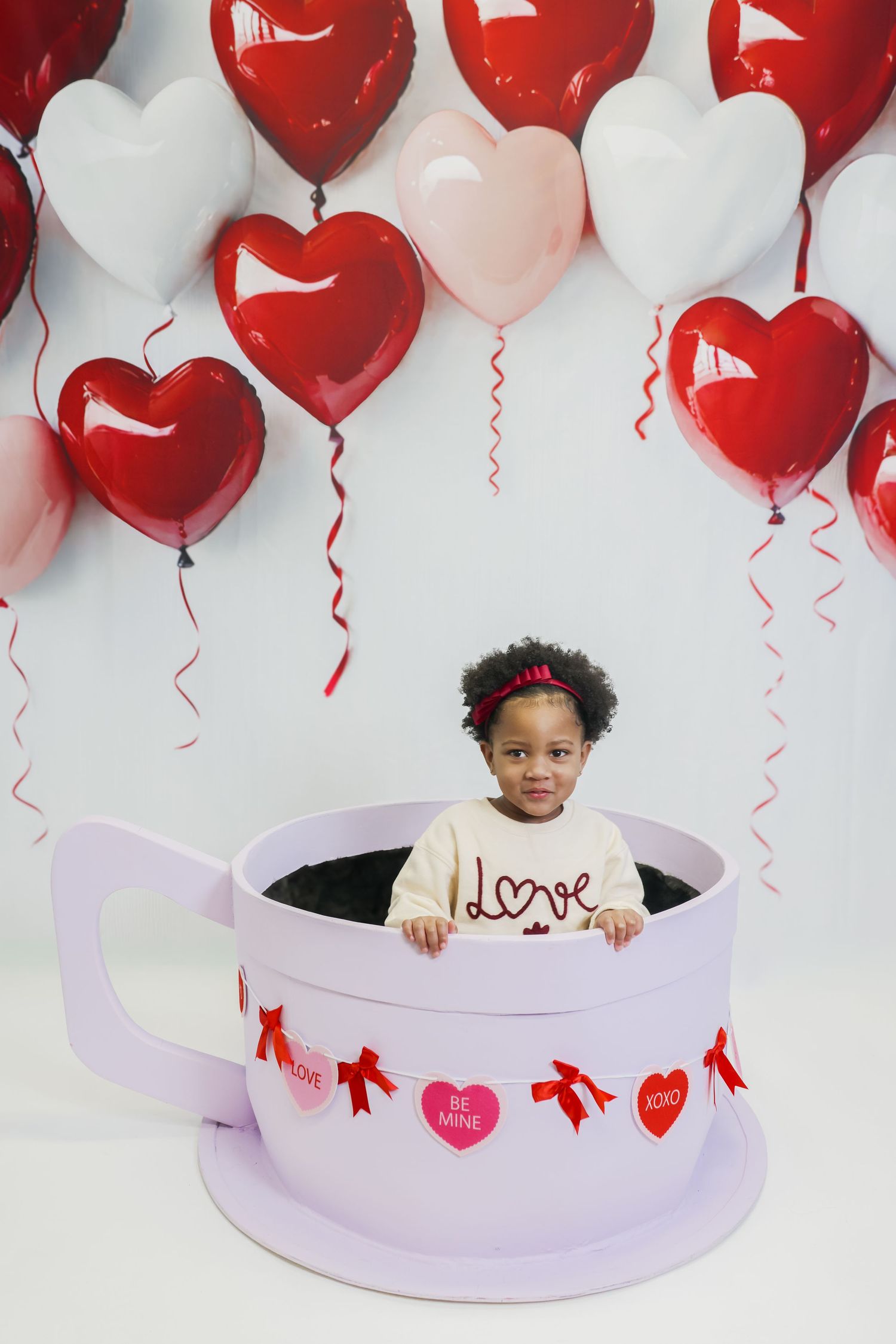 Toddler girl sitting in giant teacup prop surrounded by red and pink heart balloons for Valentine's Day photo.